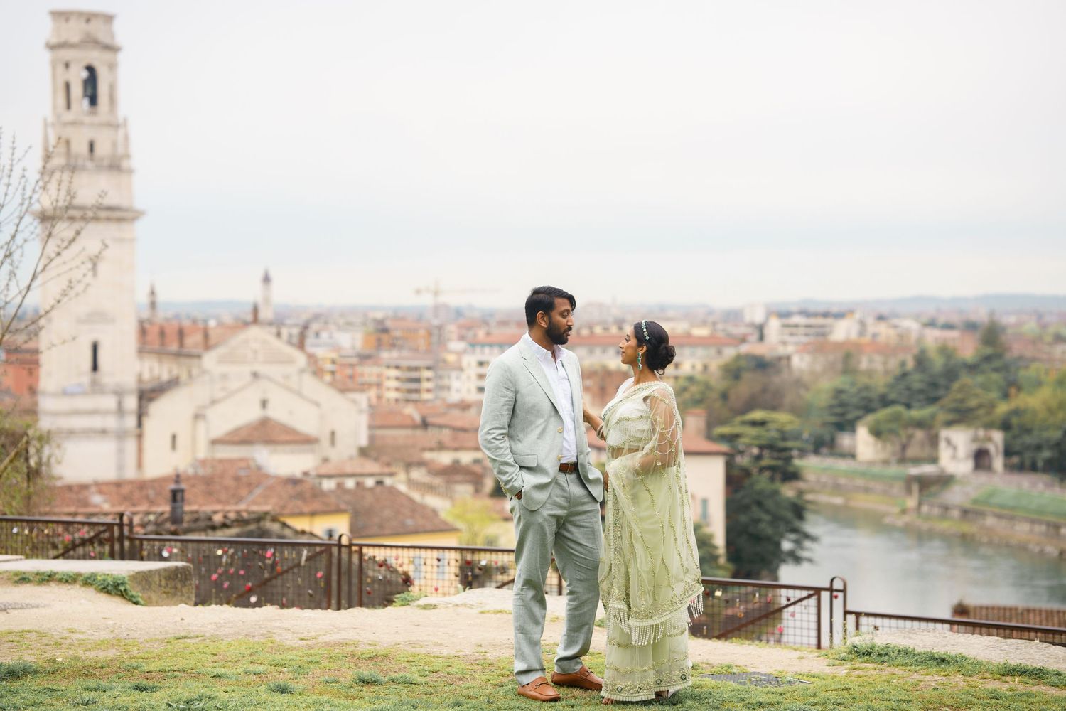 A couple in mint green outfits walks along a scenic European cityscape overlooking historic architecture and a river.