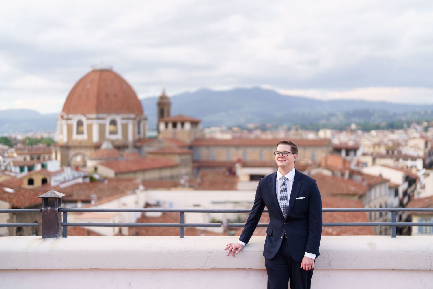 A person in a navy suit stands on a rooftop terrace overlooking Florence, Italy with the Duomo dome visible in the background.