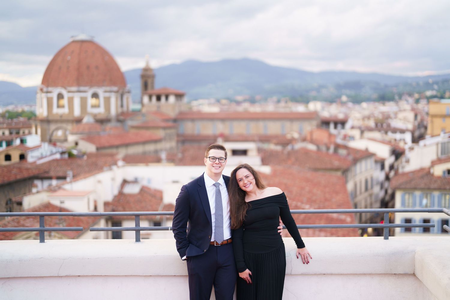 Couple enjoying scenic views of Florence from rooftop terrace with historic red-tiled buildings and mountains in background.
