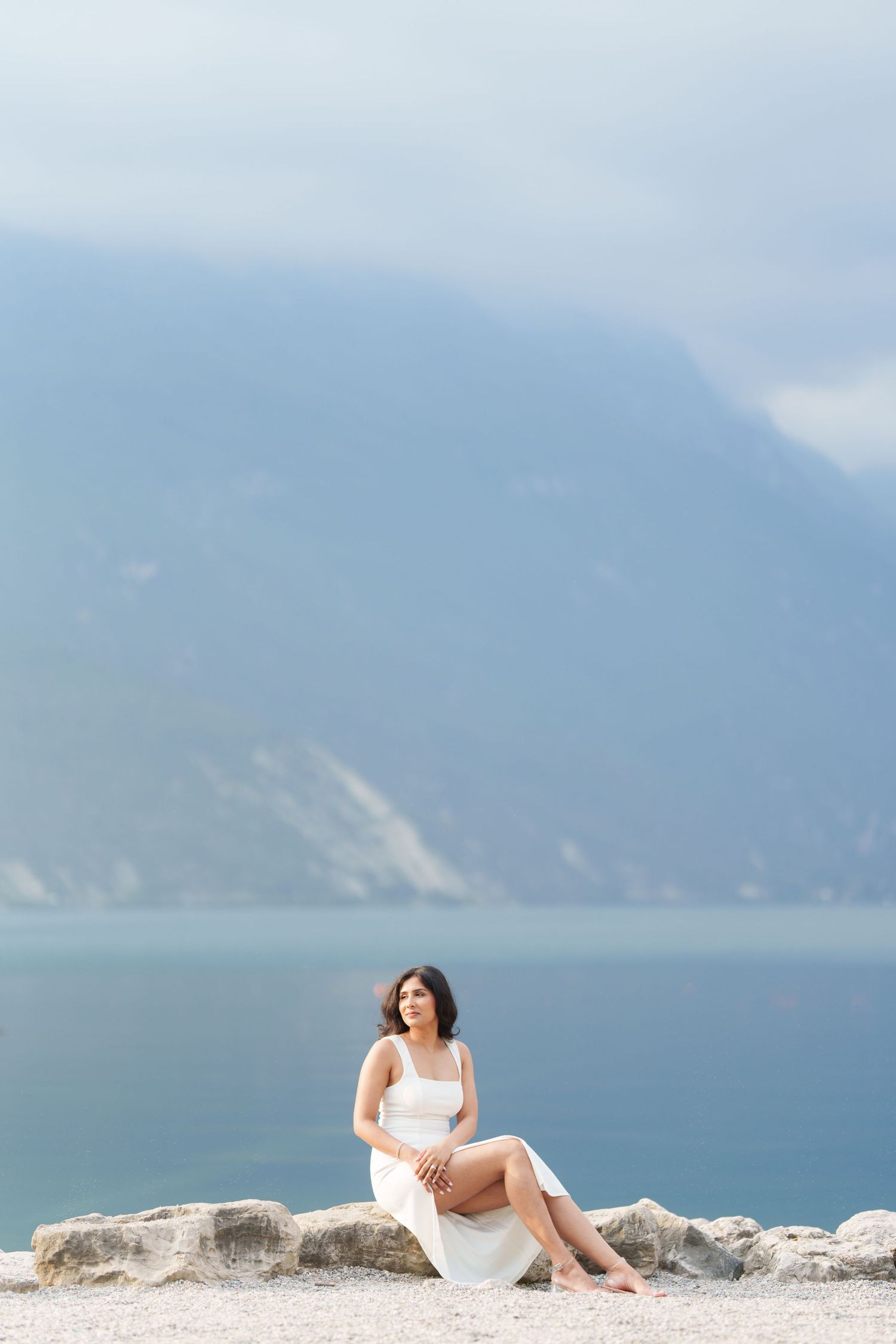 A person in a white dress reclines on a sandy beach with misty mountains and ocean in the background.