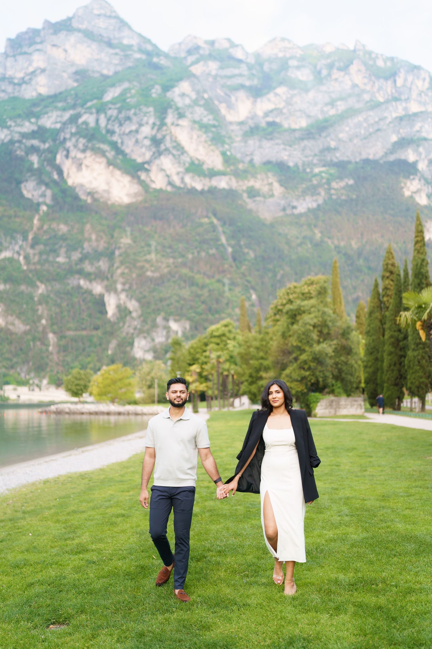 A couple walks hand in hand across a lush green lawn with majestic mountain peaks rising in the background.