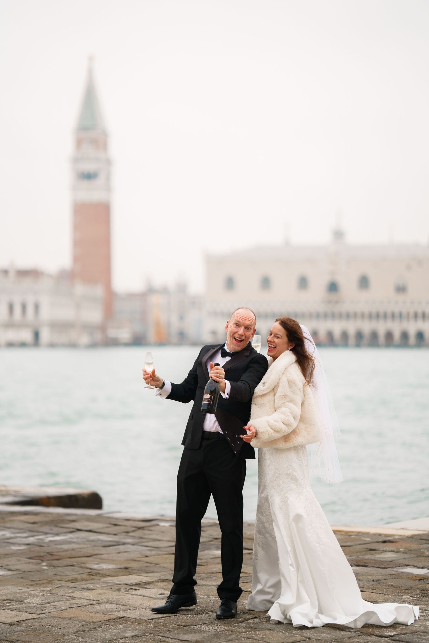 A couple in wedding attire sits by the water with Venice's iconic St Mark's Campanile in the background.