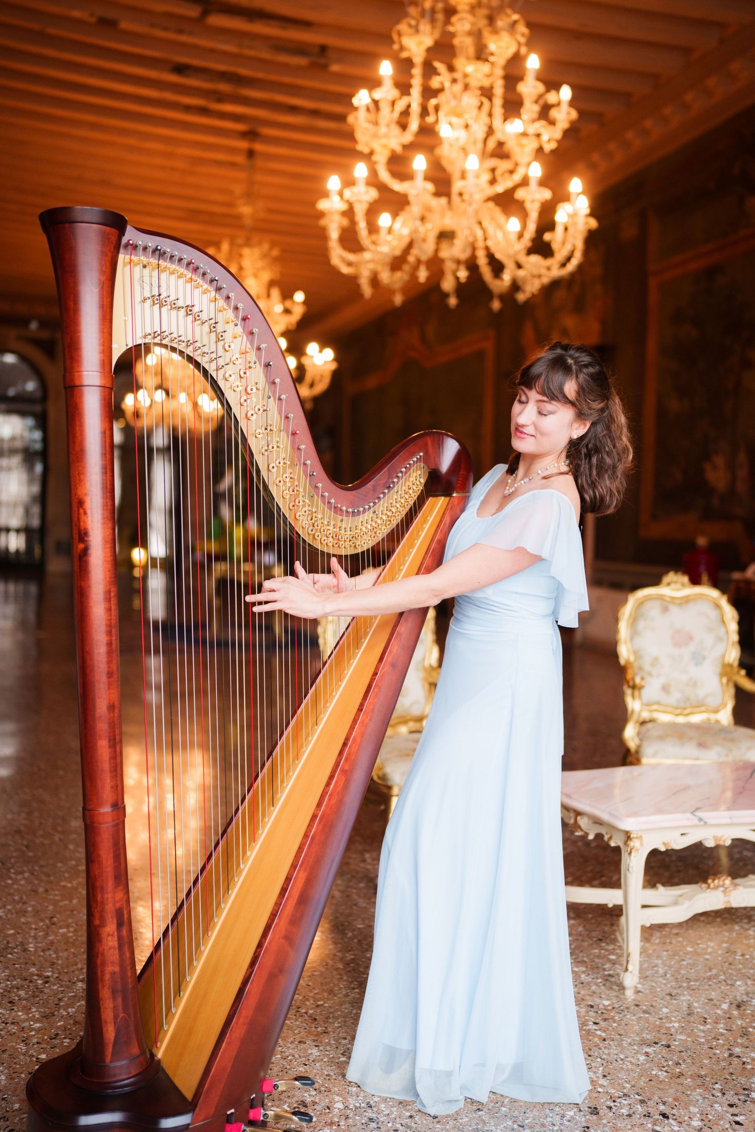 A harpist in a flowing white dress performs in an elegant room with crystal chandeliers.