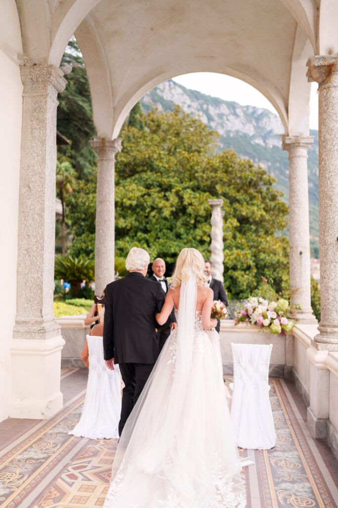 Wedding ceremony procession along a covered colonnade with scenic mountain backdrop.