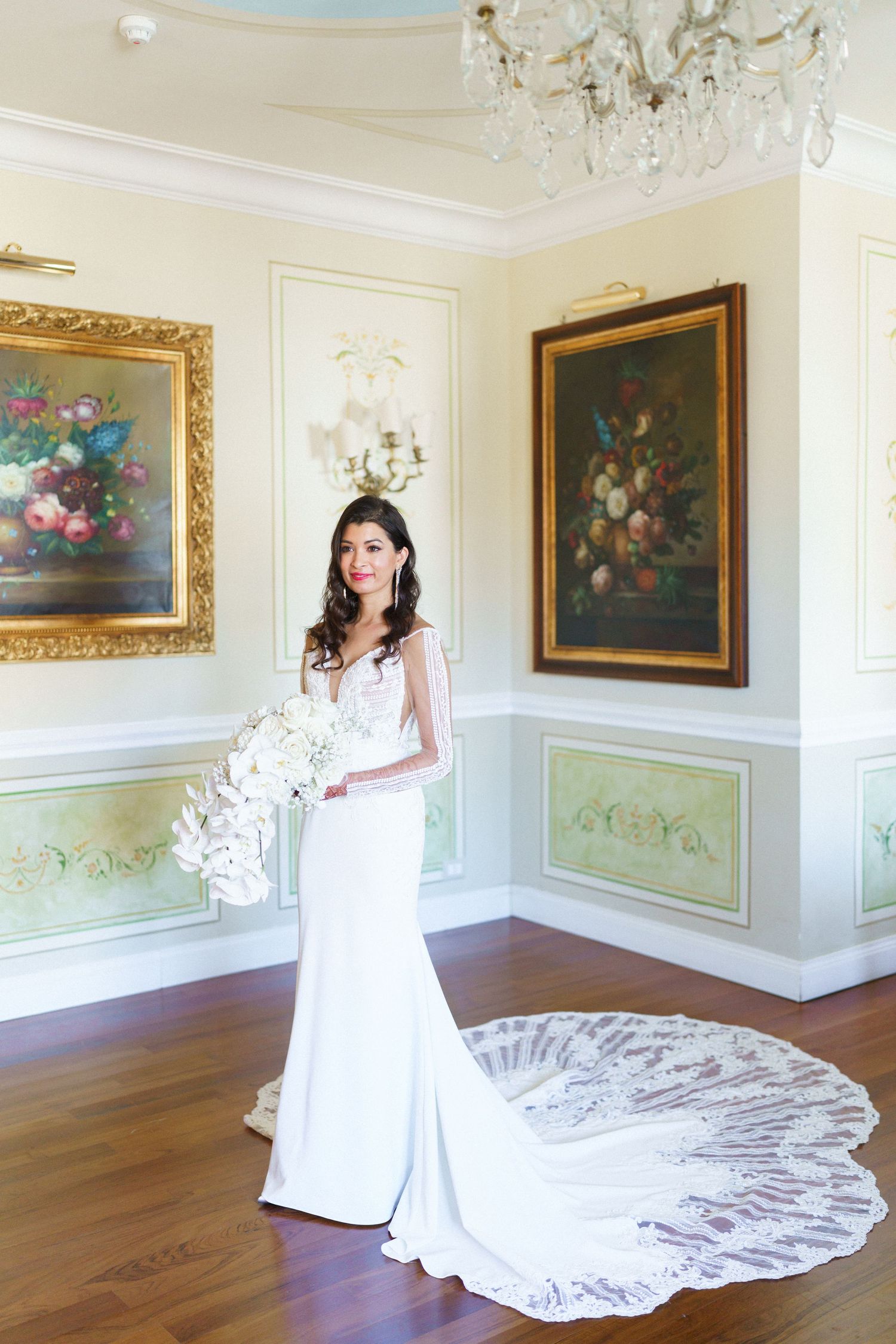 Elegant bridal portrait in white gown with long train in ornate room with floral paintings.