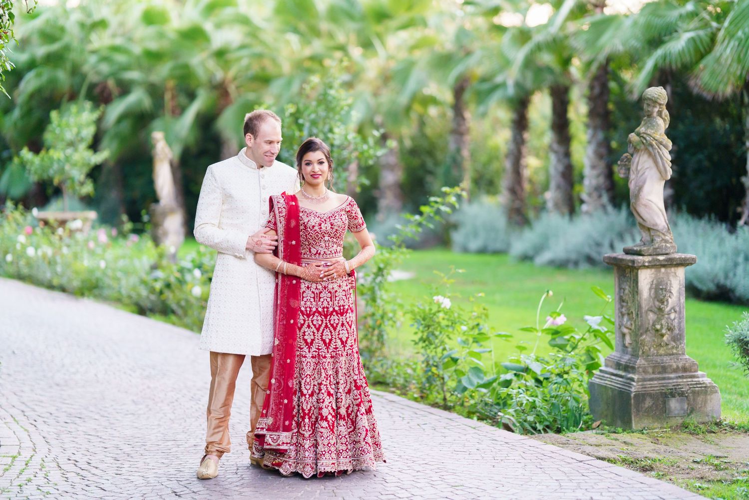 A couple walks together through a garden path lined with palm trees and stone sculptures.