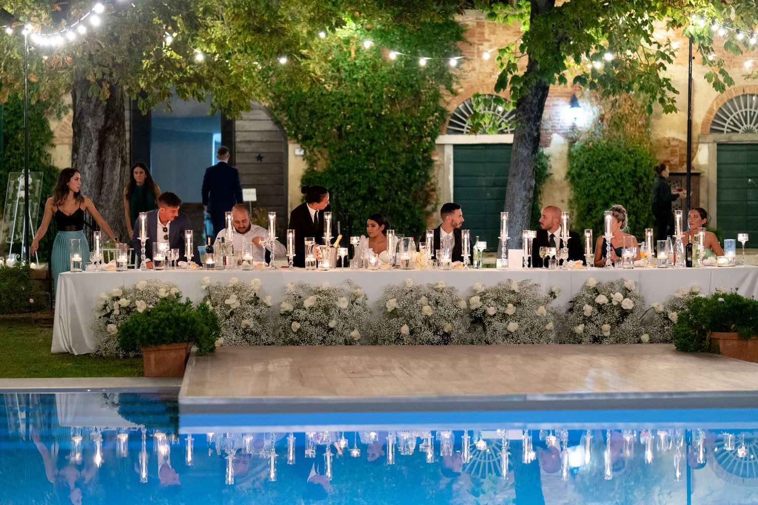 Long banquet tables set with candles and floral arrangements beside a pool under string lights.