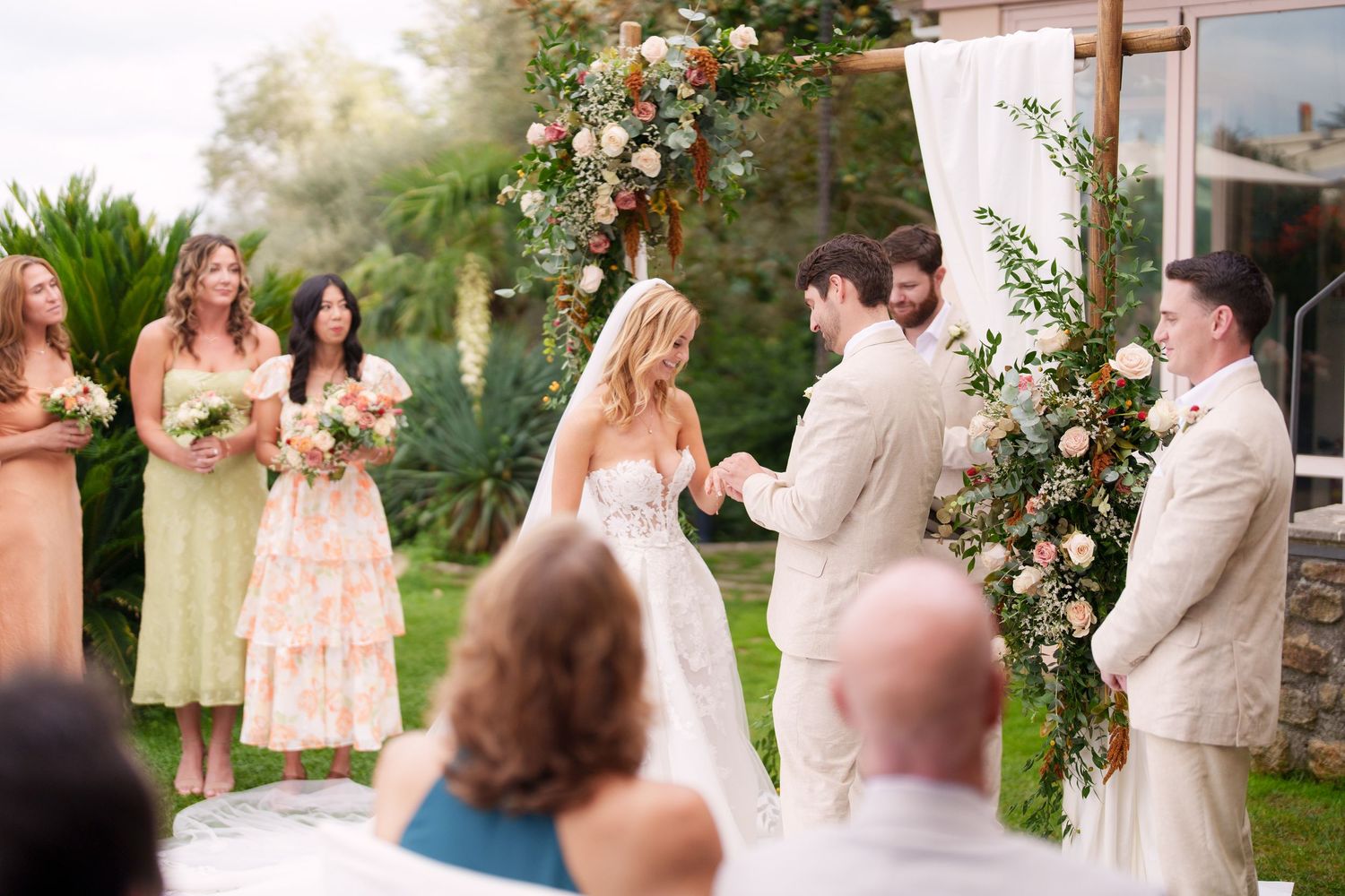 Wedding ceremony in progress under a floral arch with guests seated on white chairs.