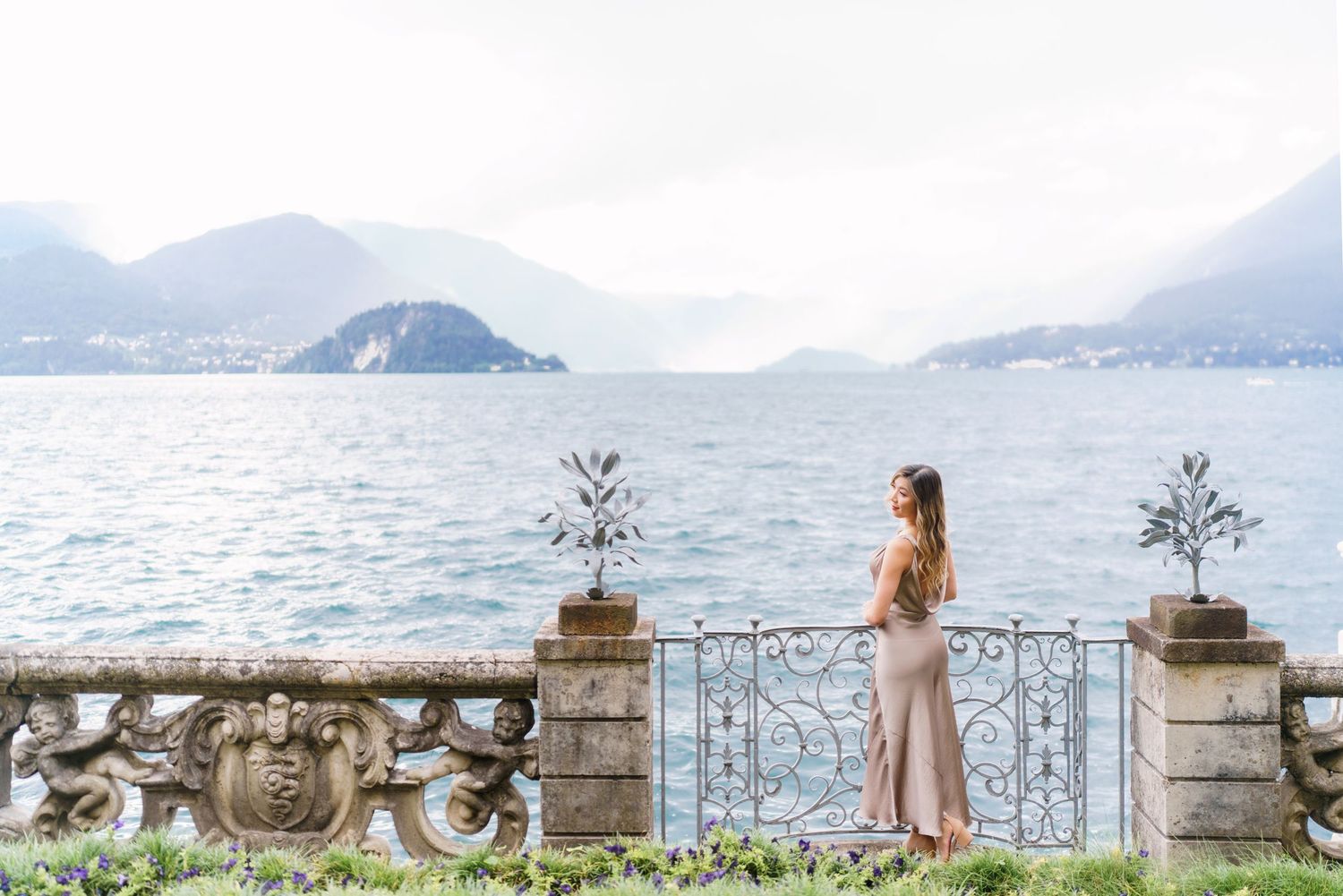 A person in a pink dress stands at an ornate stone balcony overlooking Lake Como and mountains in Italy.