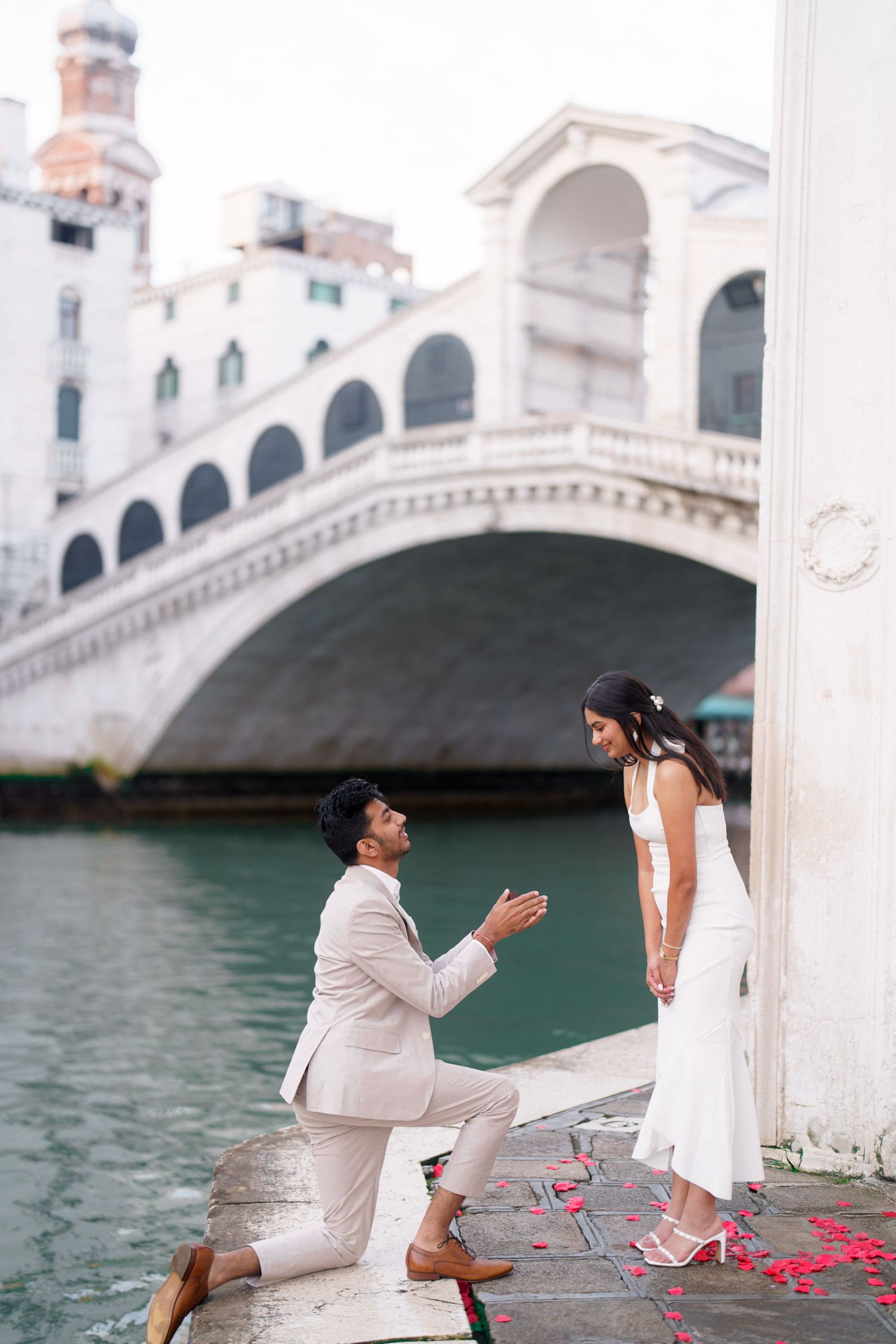 A romantic marriage proposal scene by a waterfront with white arches and rose petals scattered on the ground.