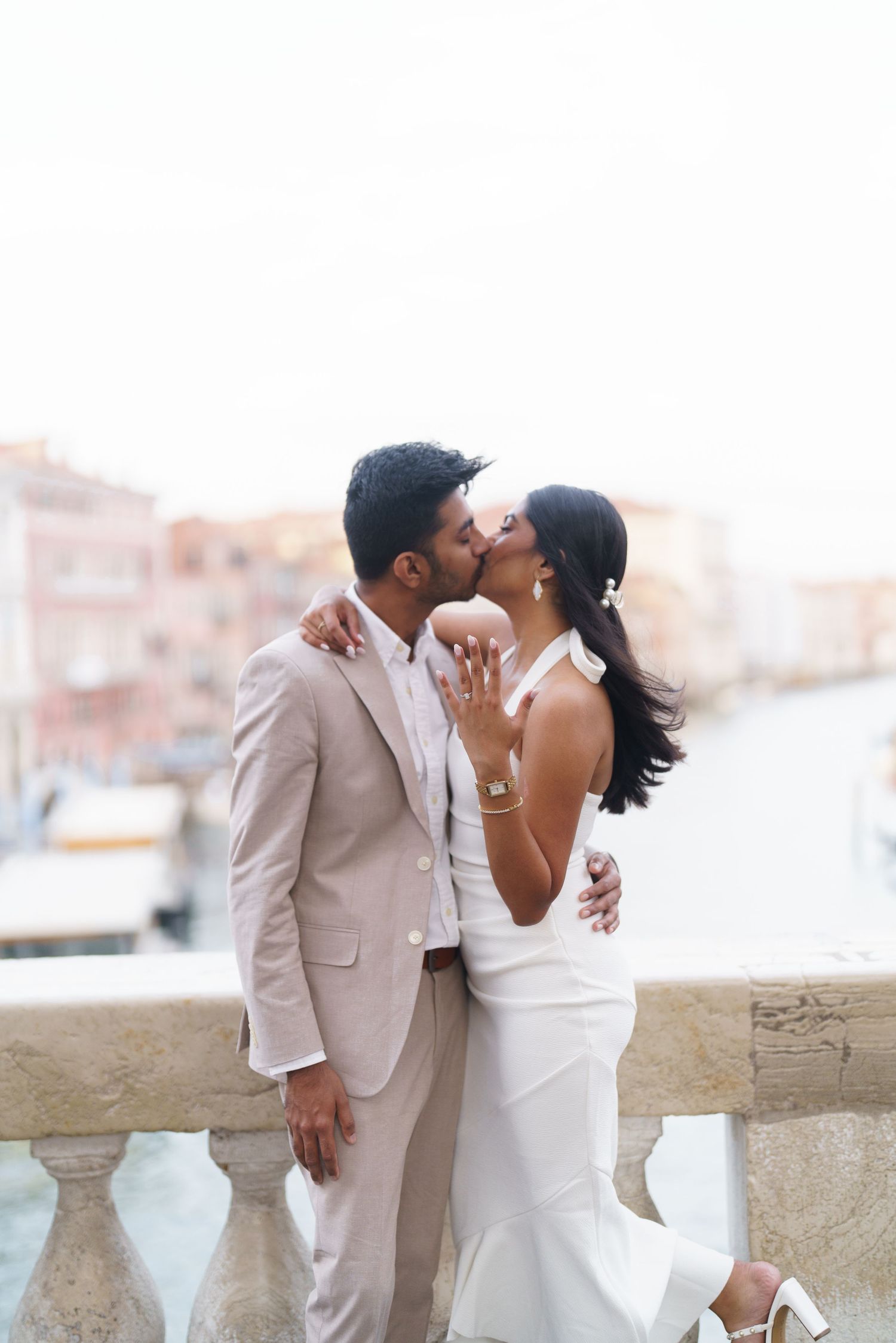 A romantic couple in elegant white attire share an intimate moment while posing against a stone balcony.