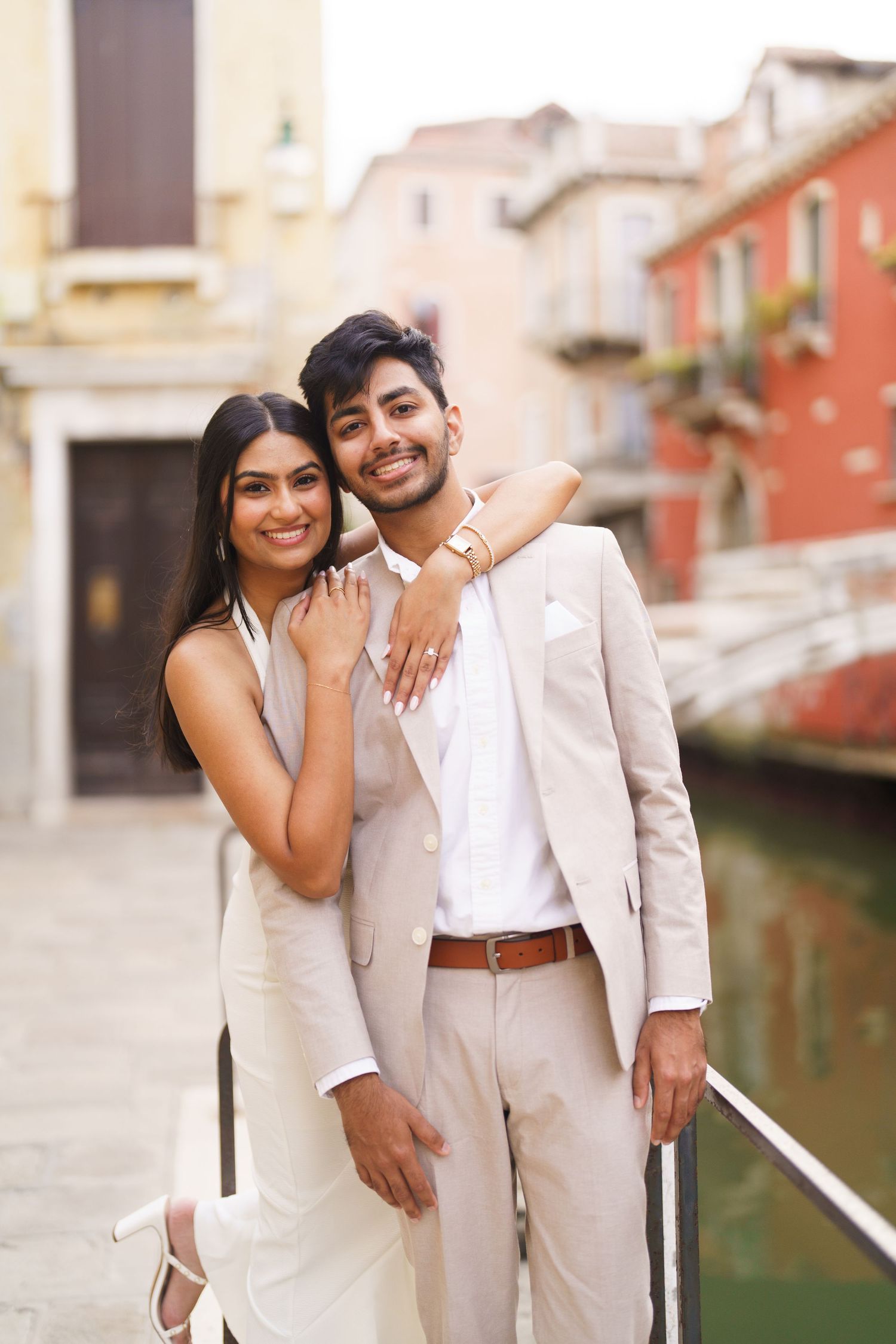 A couple in coordinated beige outfits embrace while standing along a canal in Venice, Italy.