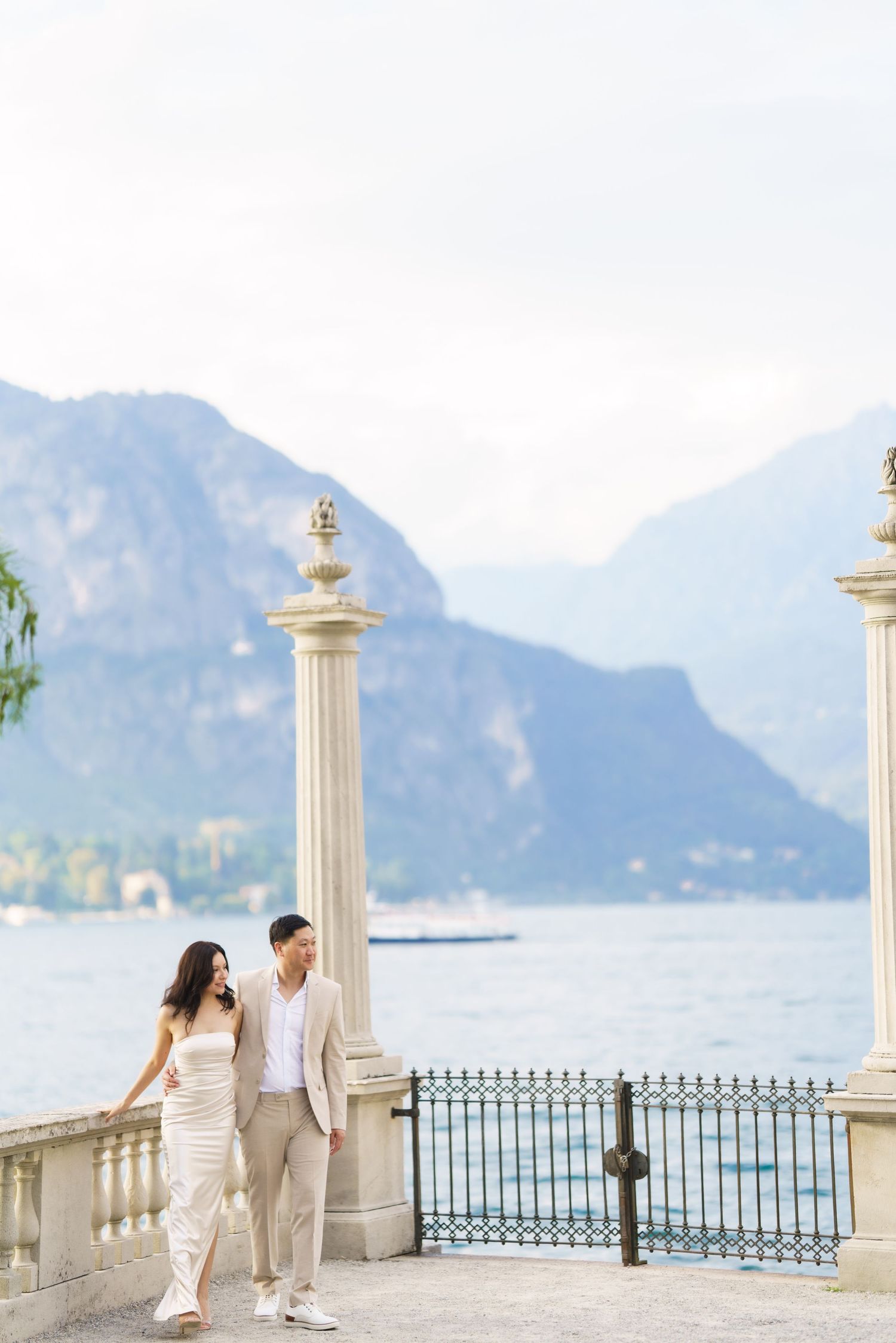 A couple walks along an elegant seaside terrace with white columns and mountain views in the background.