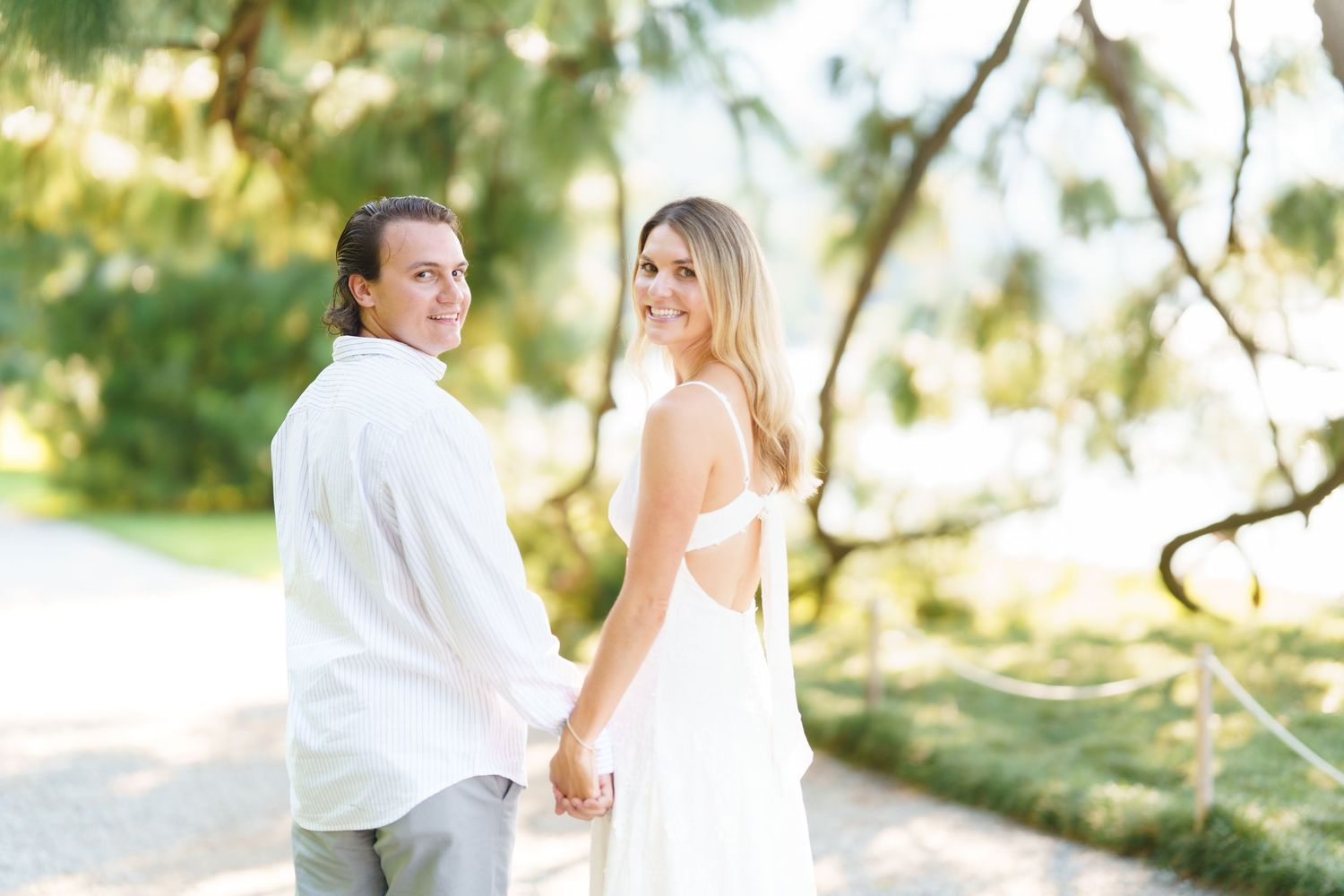 Couple in white outfits poses together in a romantic outdoor photoshoot with sunlit trees in the background.