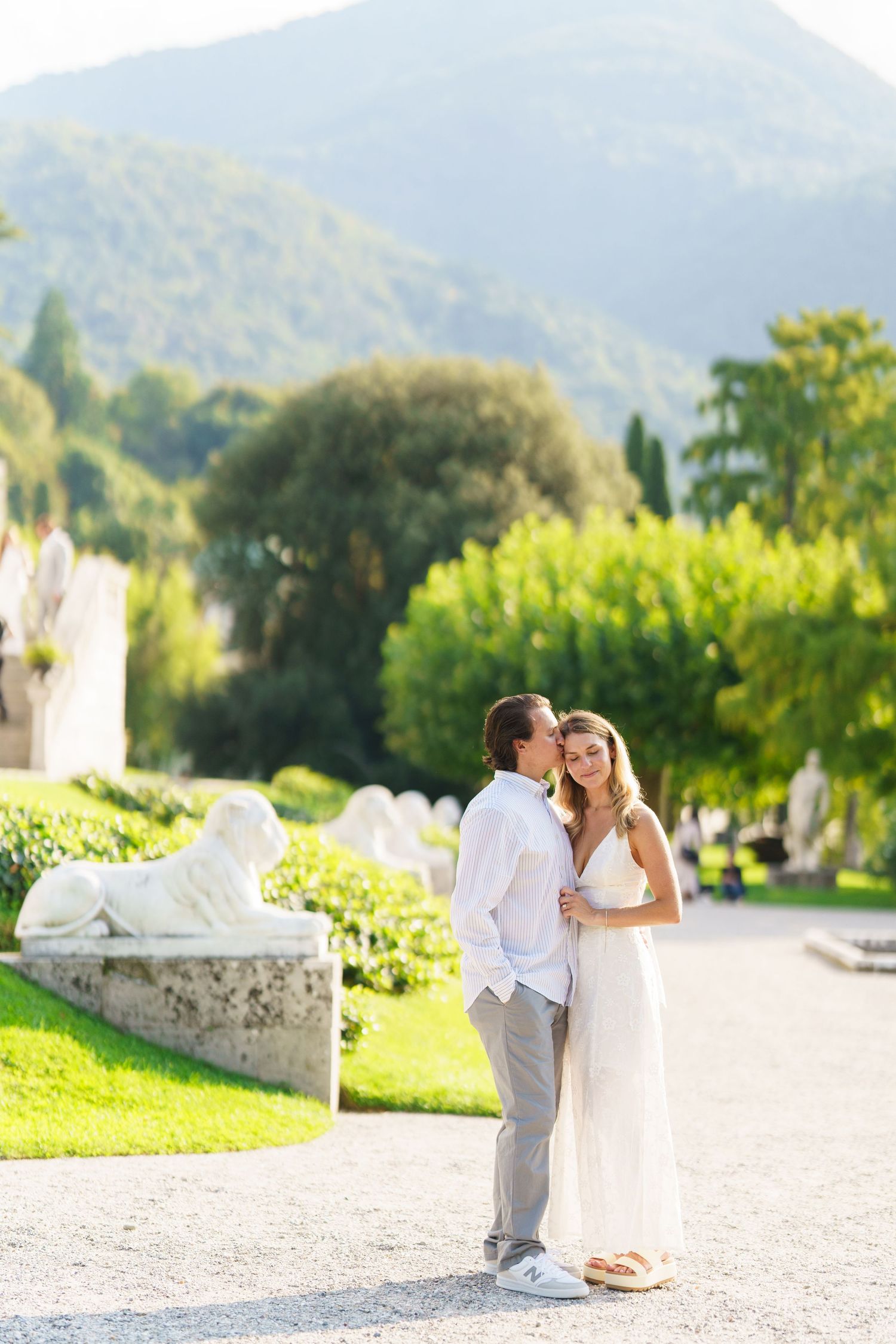 A romantic couple shares tender moments in a sunlit garden with mountain views and white stone architecture.