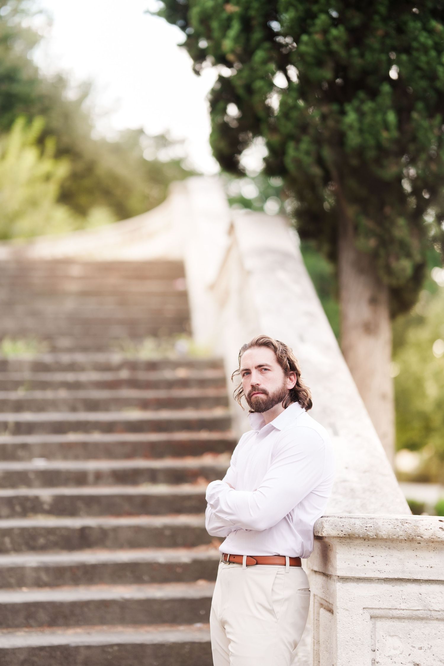 A person in a white shirt and brown belt sits thoughtfully on stone steps in an outdoor garden setting.