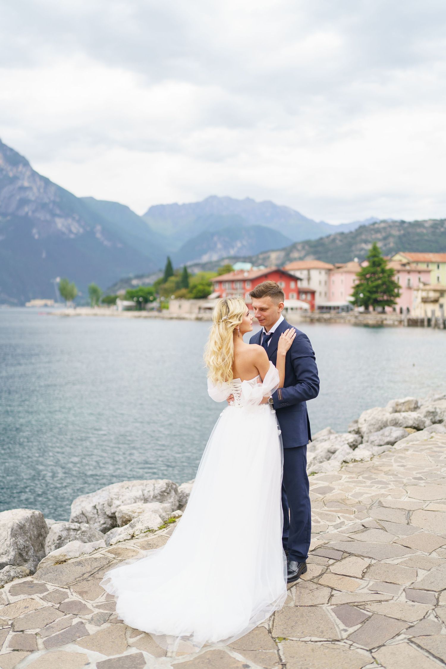 Romantic couple embraces on a stone walkway overlooking Lake Como with mountains and Italian villas in the background.
