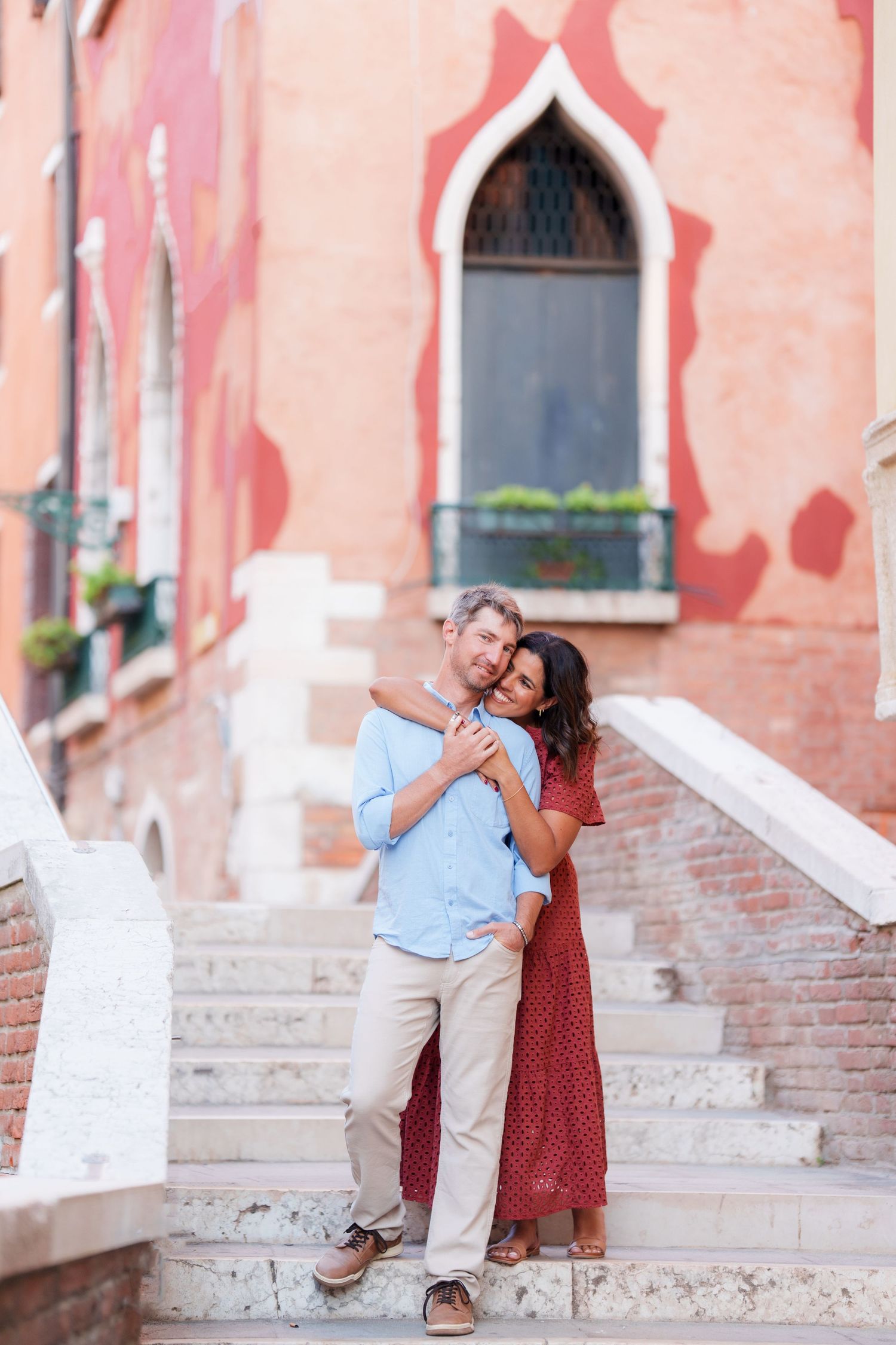 Couple embracing on stone steps in Venice, Italy, with a red building and arched window in the background.