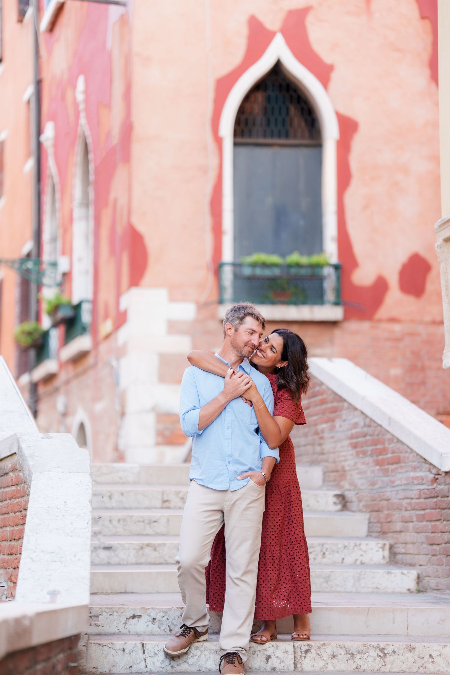 Couple embracing on marble steps in Venice, Italy, with a rustic red building and Gothic window backdrop.
