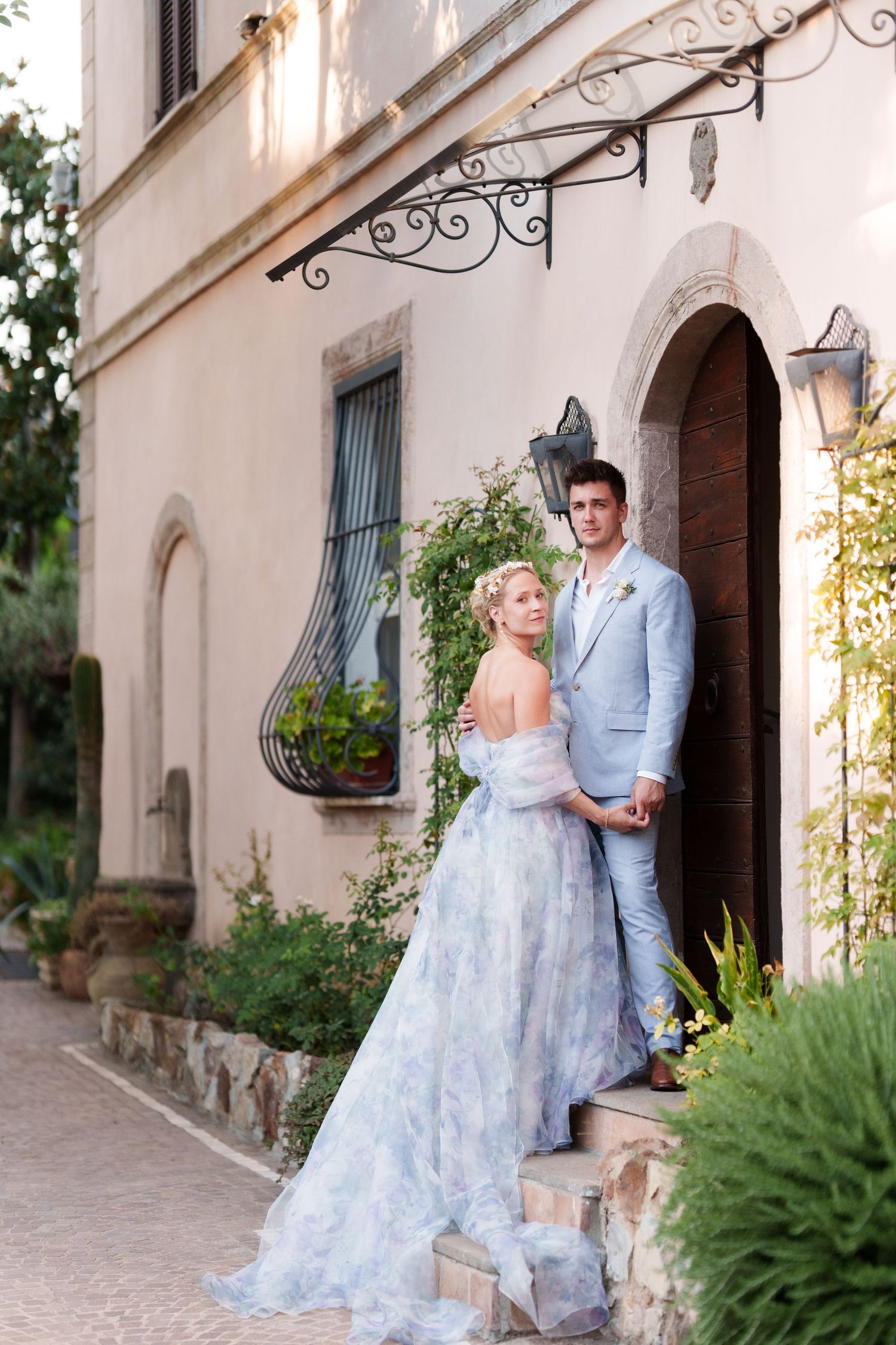 Bride in floral gown and groom in blue suit pose outside an elegant Mediterranean villa surrounded by lush greenery.