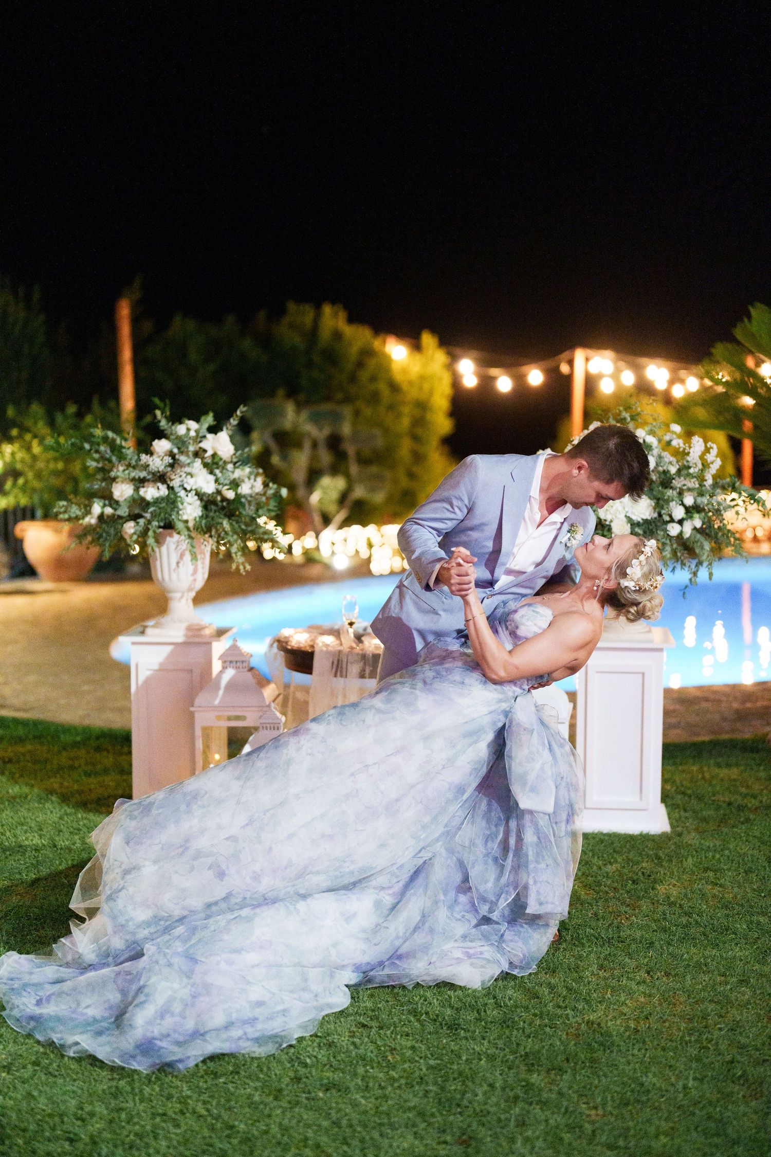 Couple sharing a romantic dip kiss in elegant wedding attire beside a pool at night with floral arrangements.