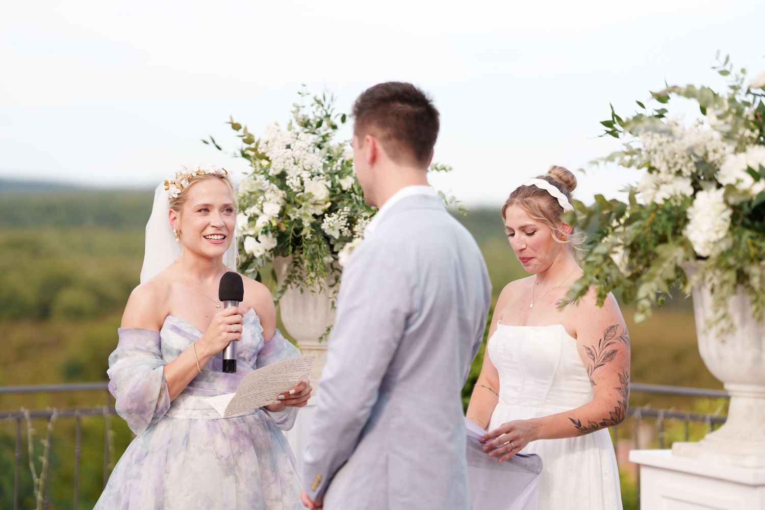 Outdoor wedding ceremony with officiant holding microphone, groom in gray suit, and bride in white dress by floral arrangements.
