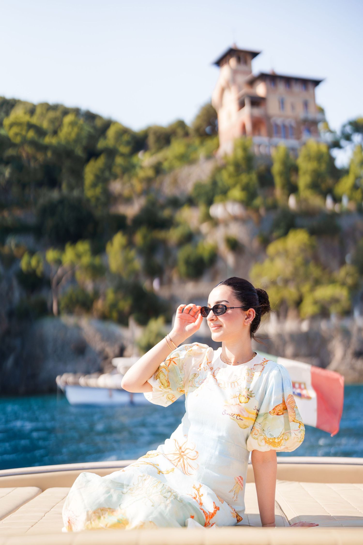 Elegant woman in floral dress adjusting sunglasses on a yacht, with a cliffside villa and blue sea in background.