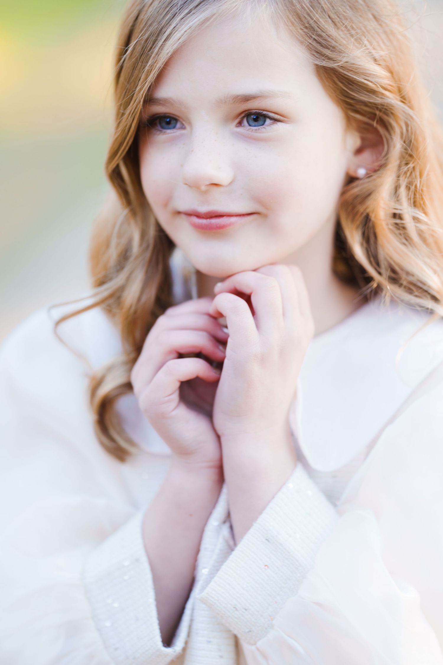 Young girl in white dress with pearl earrings, hands clasped near chin, smiling softly outdoors.