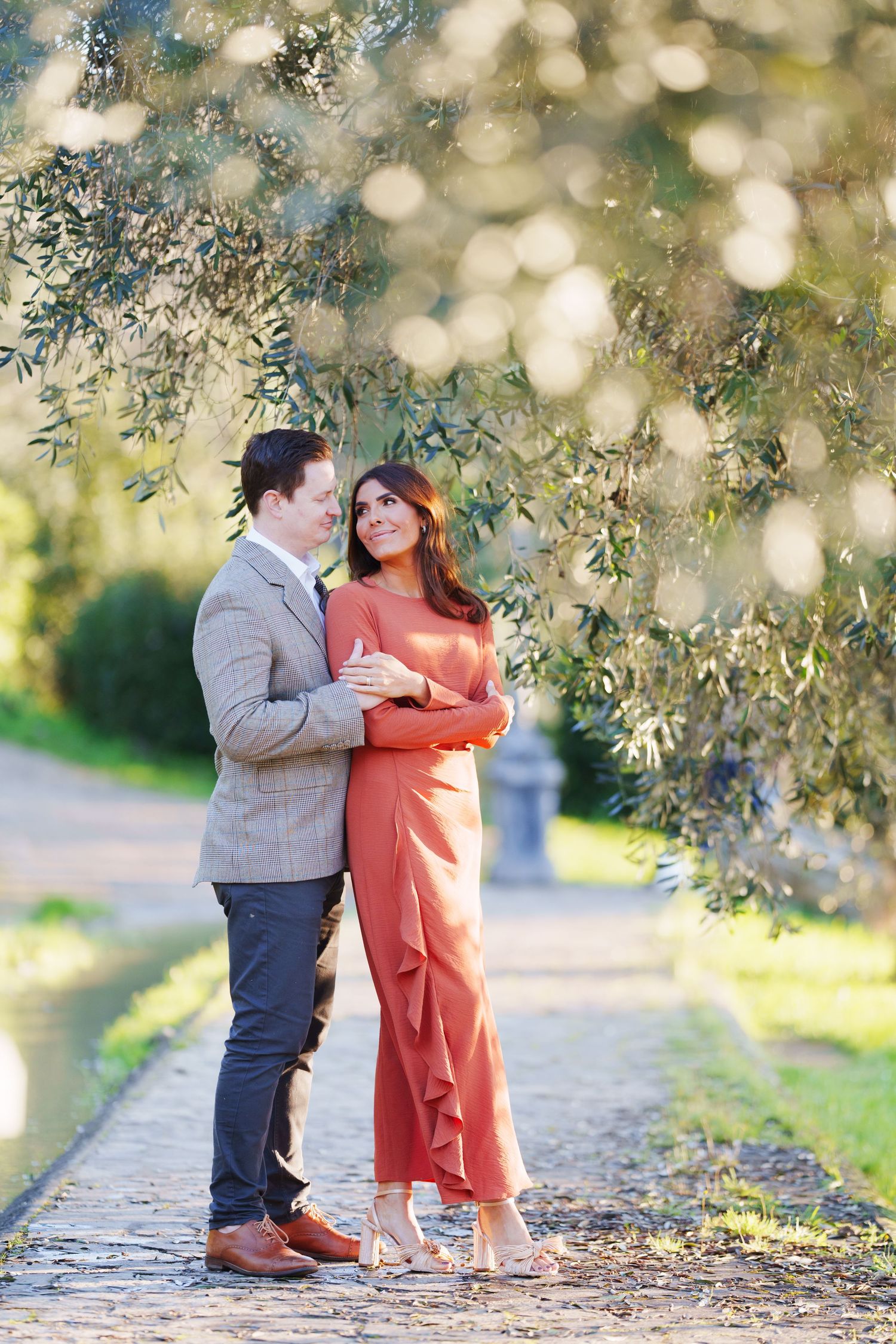 Couple embracing outdoors under olive trees on a stone path, woman in rust jumpsuit and man in gray suit.