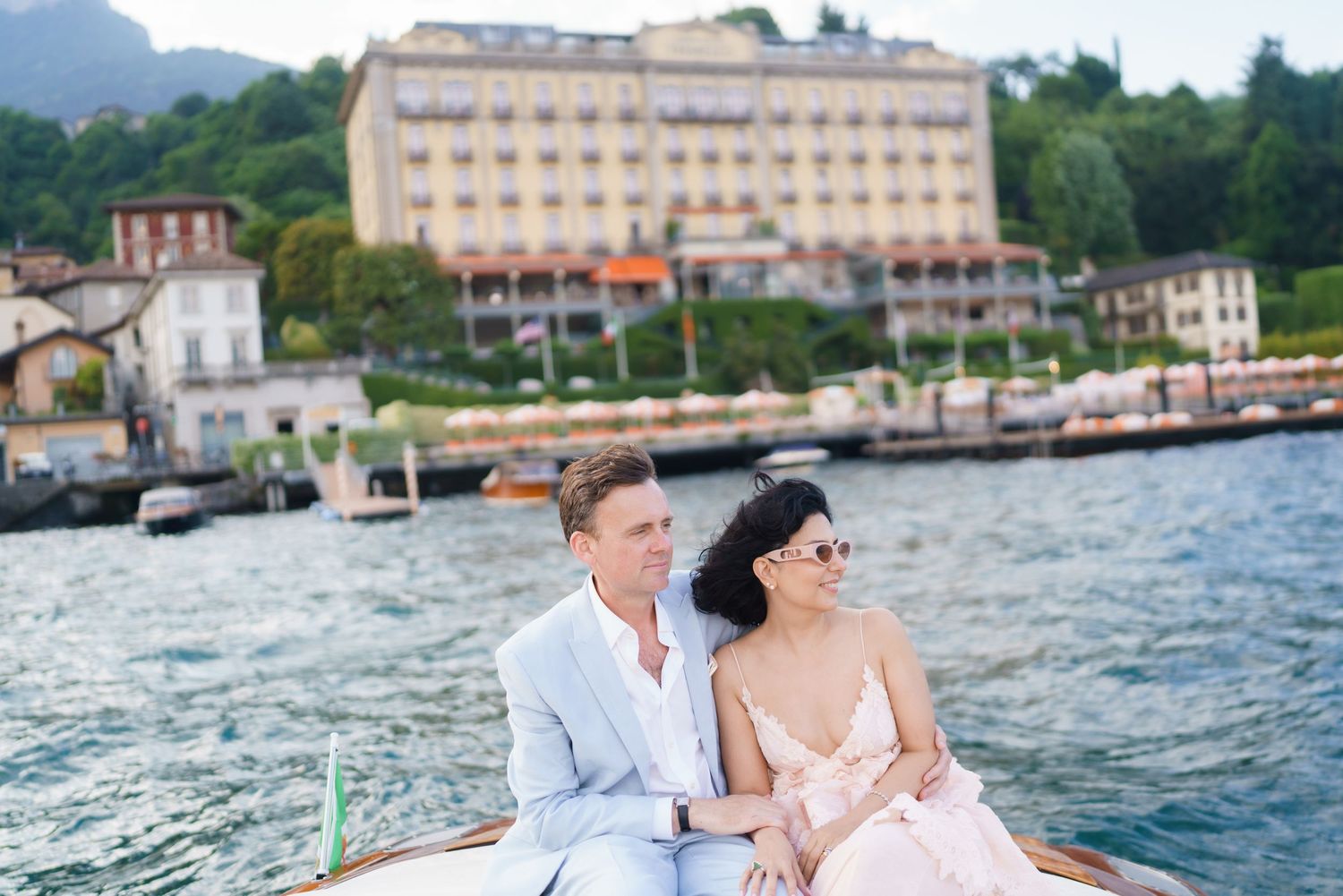 Couple sitting on a wooden boat on Lake Como, Italy, with a grand lakeside hotel in the background.