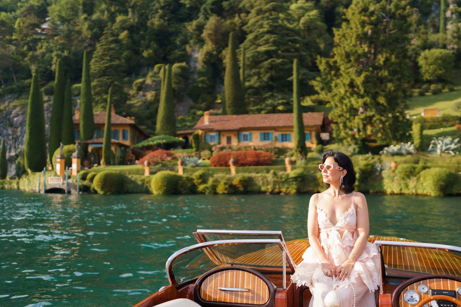 Woman in pink dress sitting on a wooden boat on turquoise Lake Como with lush hillside villa in background.