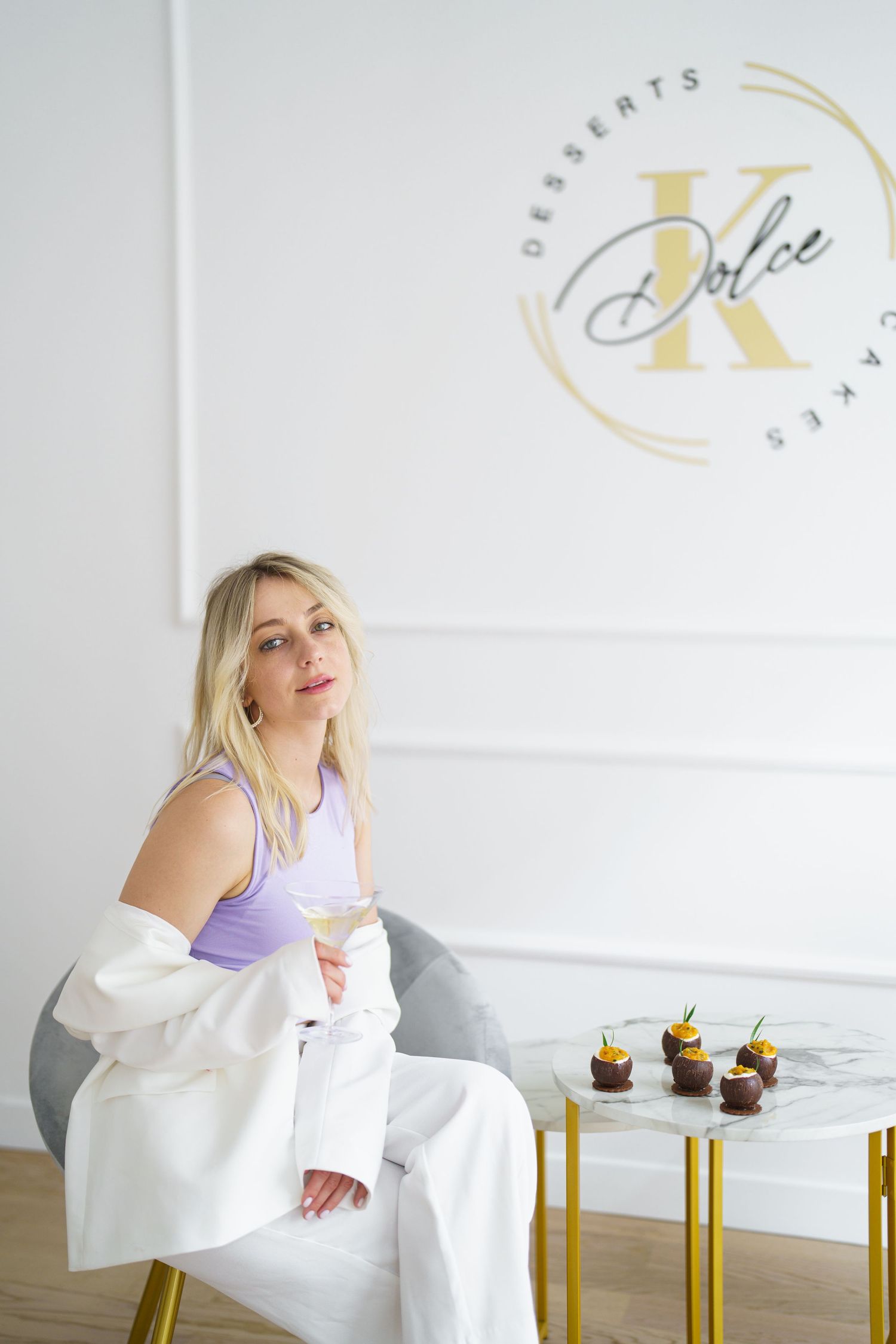 Blonde woman in white outfit sitting in a gray chair at Dolce K Desserts and Cakes bakery with chocolate cupcakes on marble table.