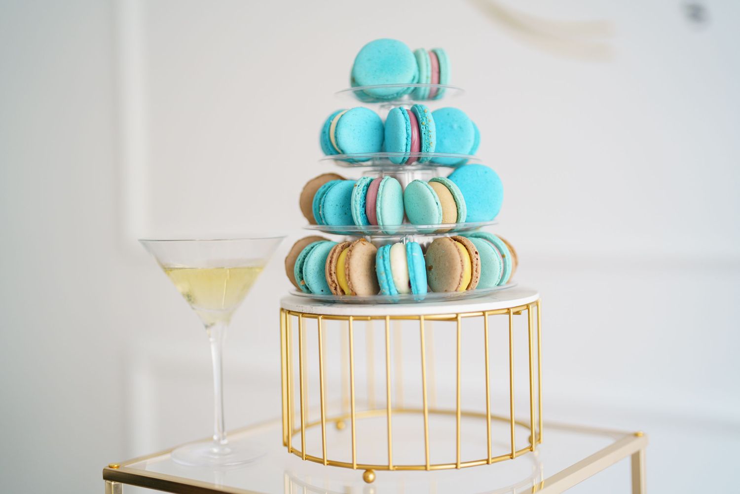 Elegant macaron tower in blue and gold tones displayed on a golden stand beside a champagne glass.