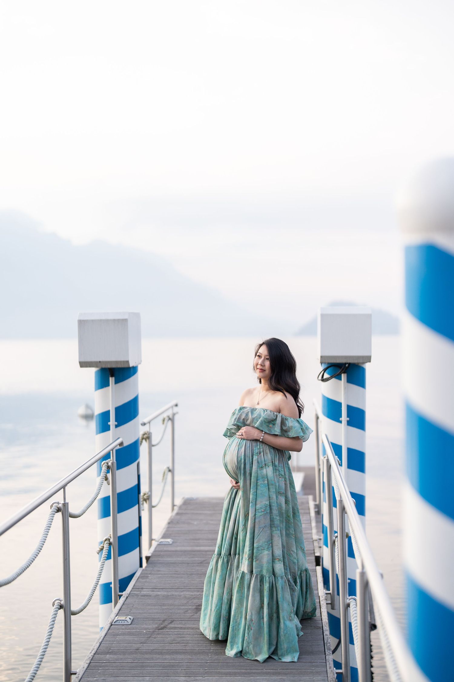 Pregnant woman in teal off-shoulder gown poses on a lakeside pier with blue striped posts and mountain views.