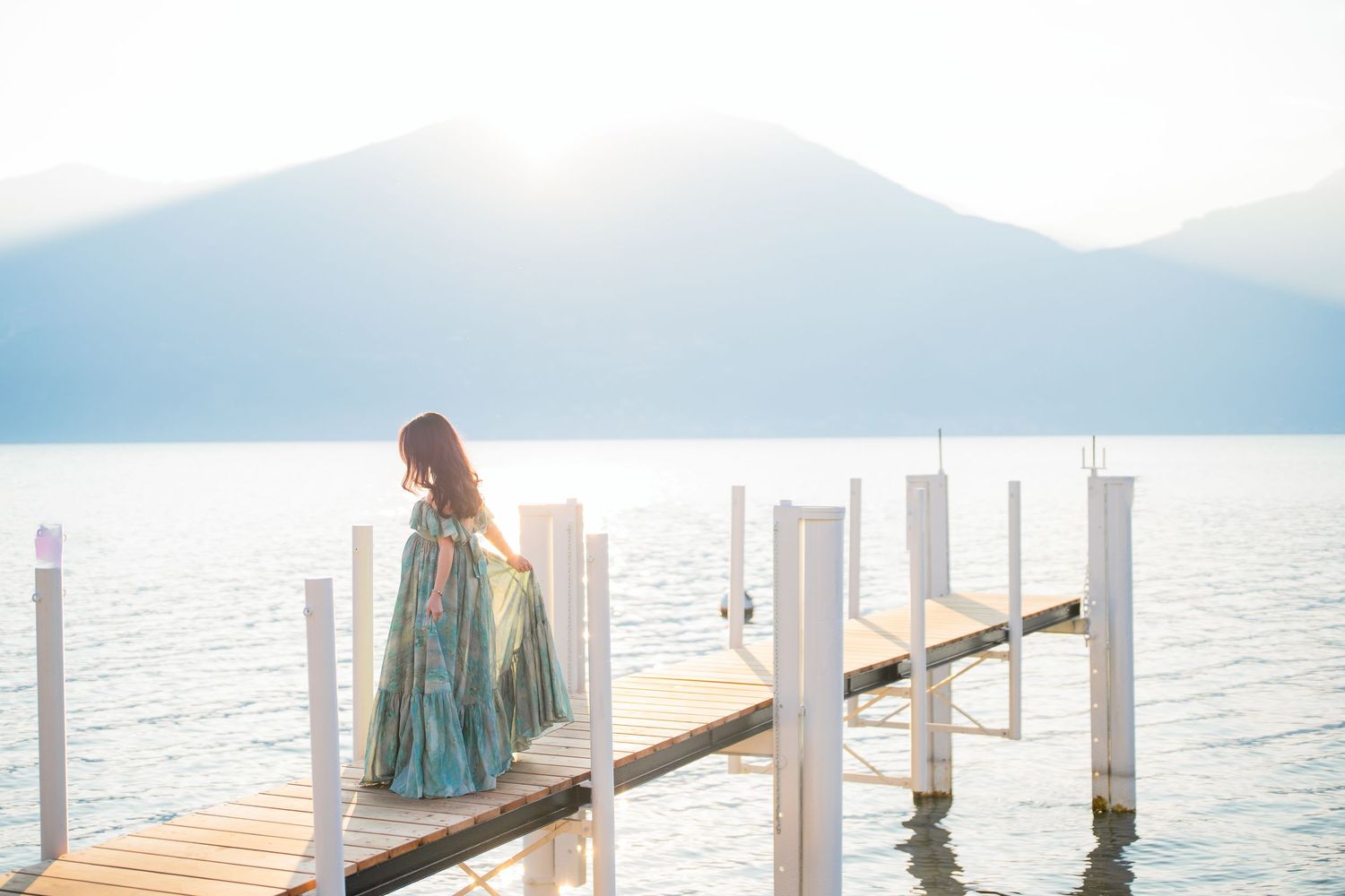 A woman in a flowing blue gown stands on a wooden dock overlooking a serene mountain lake in soft sunlight.