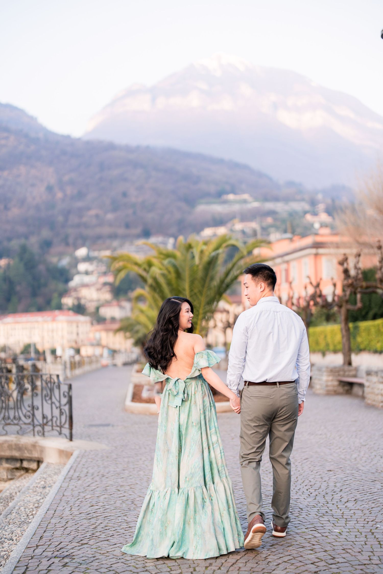 Couple holding hands on cobblestone path with mountain backdrop, woman in elegant green off-shoulder gown.