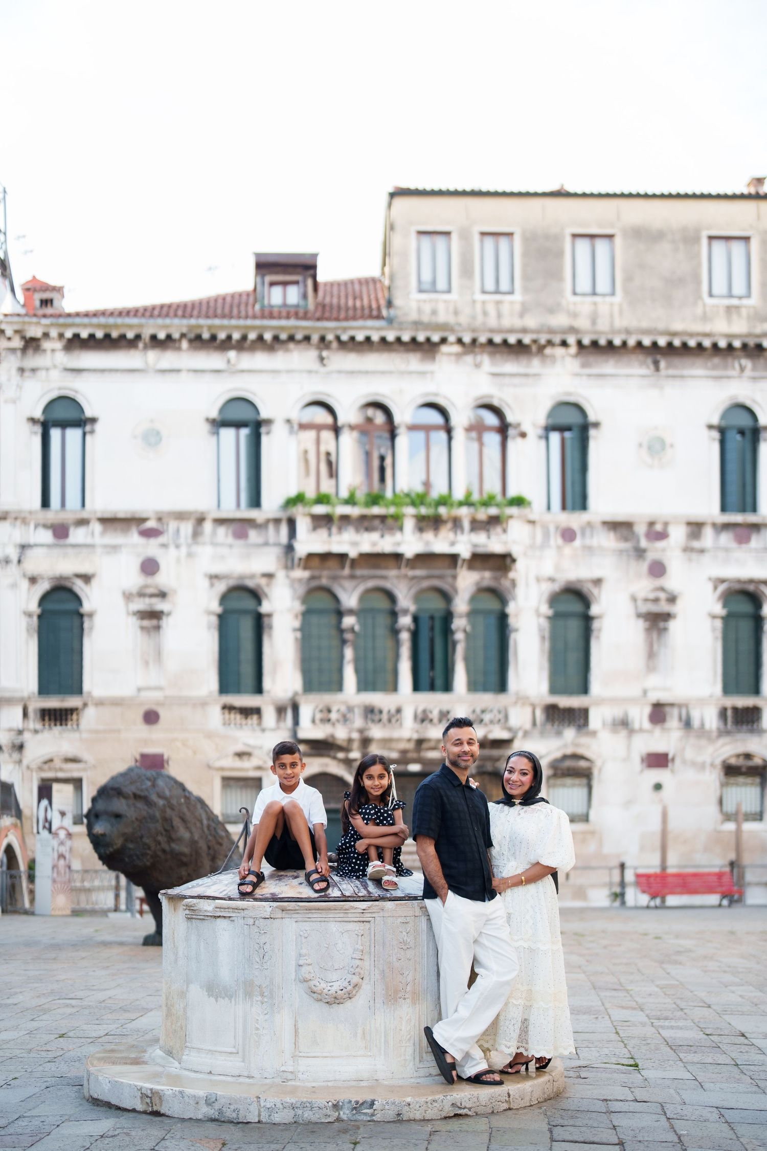 Family of four posing by a stone well in a Venice piazza, with historic Italian architecture in the background.