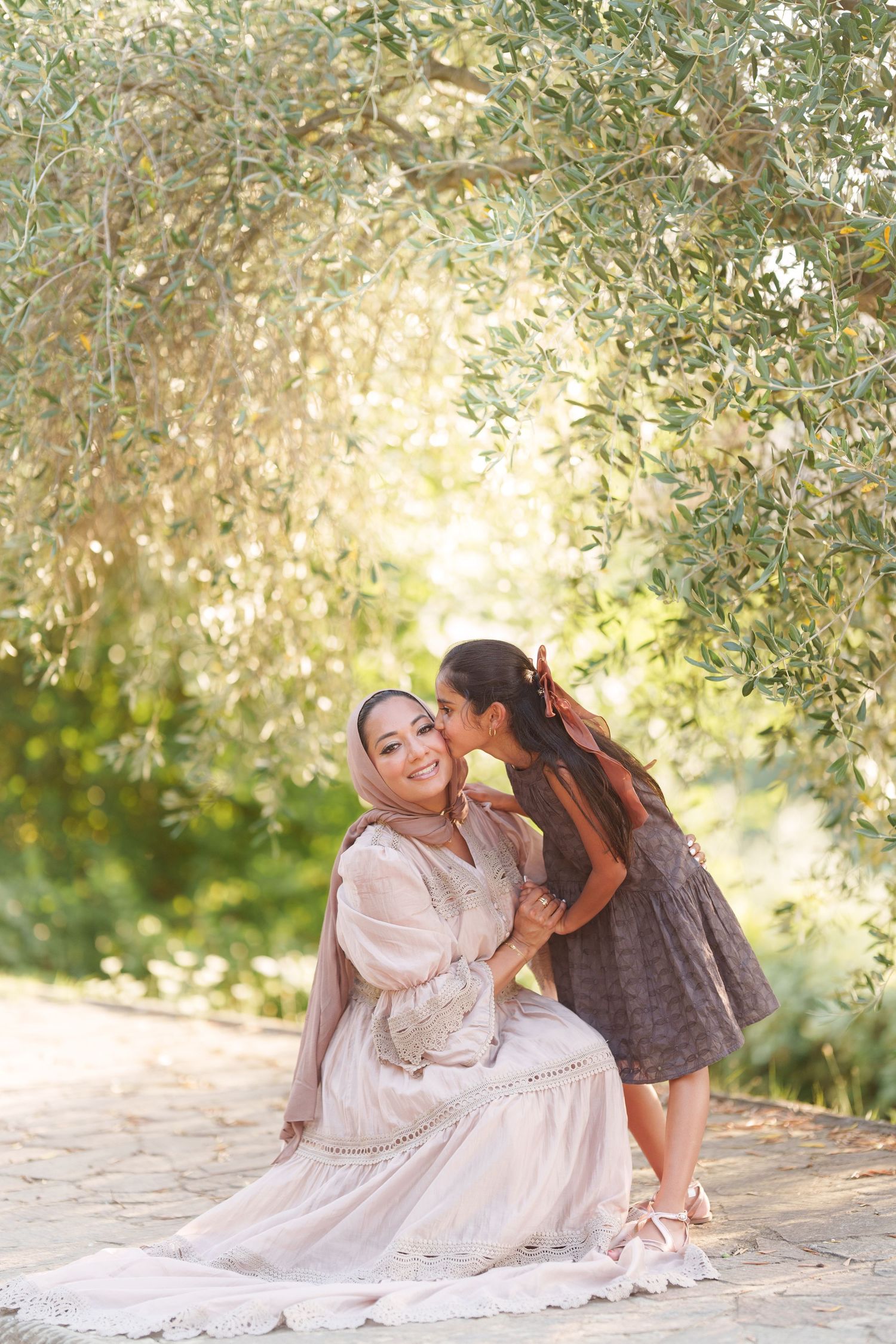 A woman in a hijab sits outdoors as a young girl kisses her cheek beneath olive trees in golden sunlight.