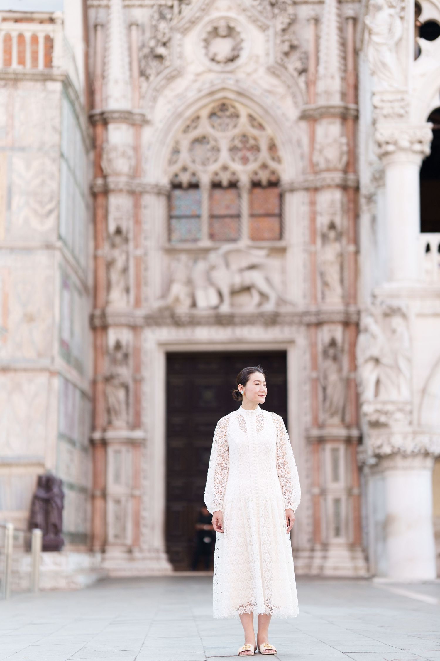Young Asian woman in white lace dress stands before ornate Gothic cathedral facade in Venice, Italy.