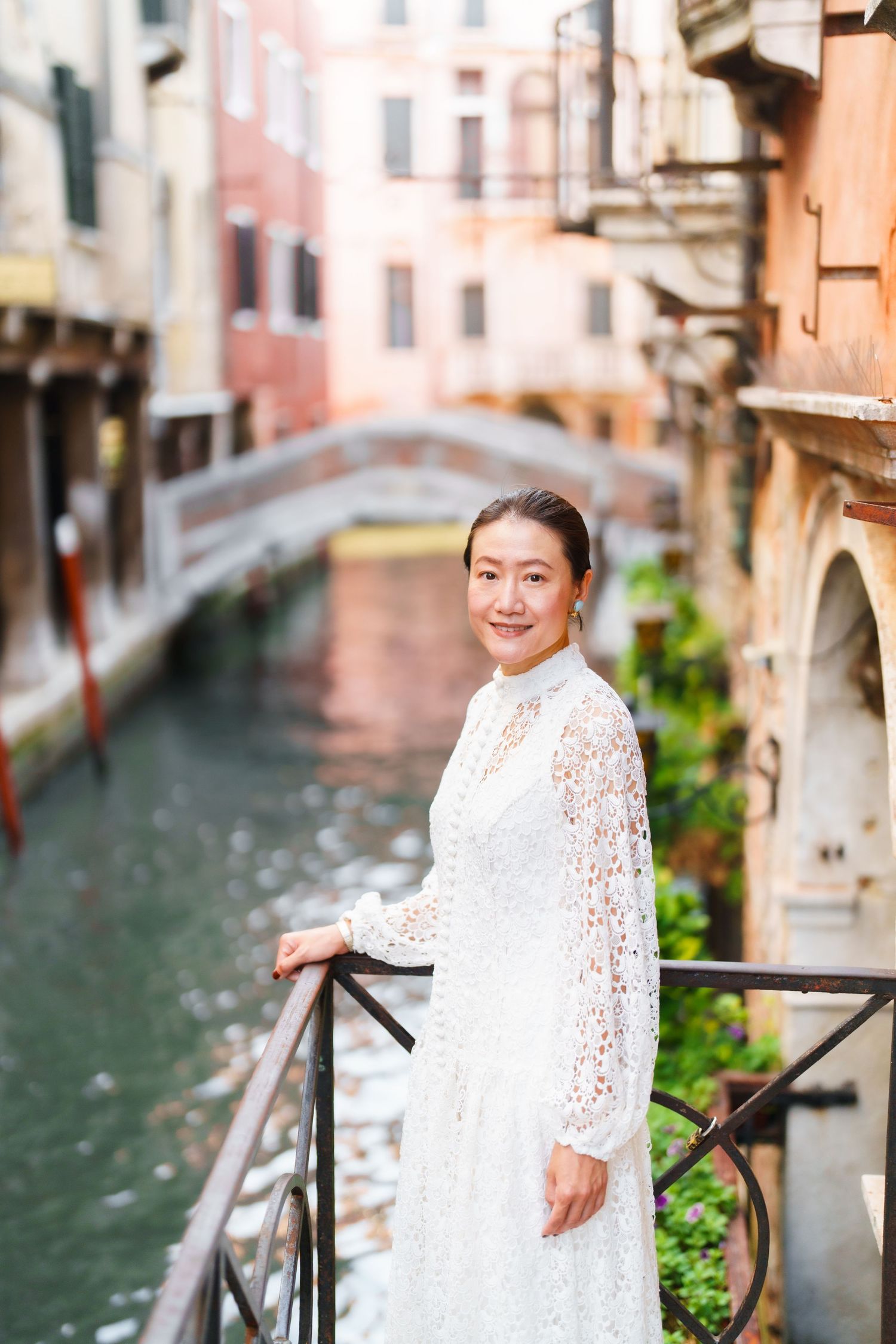 Woman in white lace dress smiling on bridge over Venice canal with historic buildings in background.