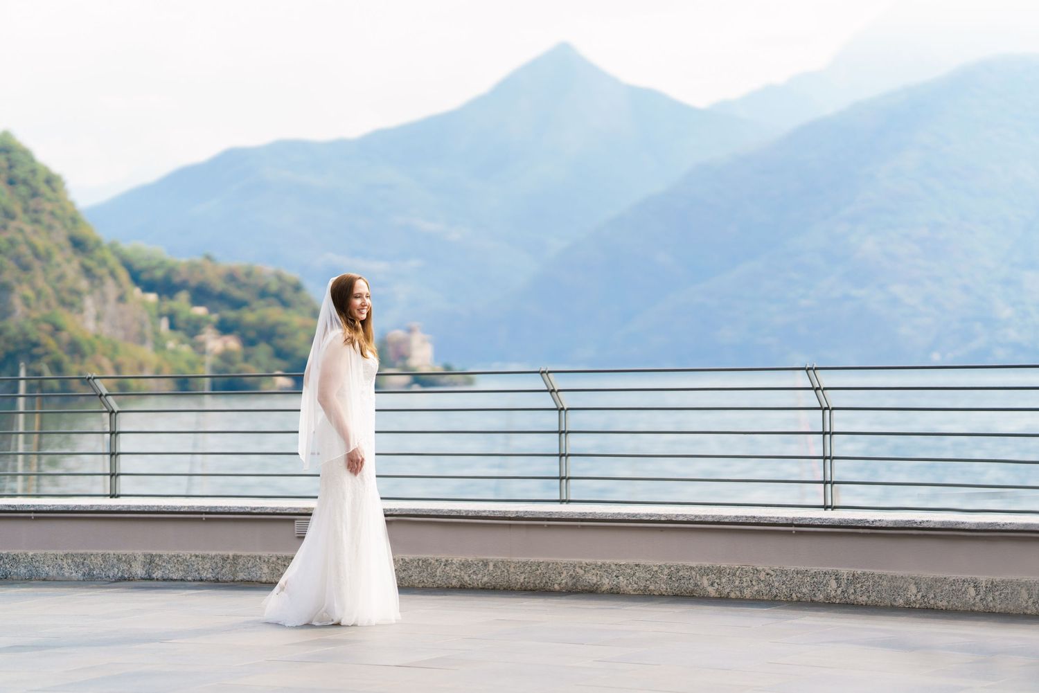 Bride in white gown and veil stands on terrace with mountain lake backdrop at Lake Como wedding.