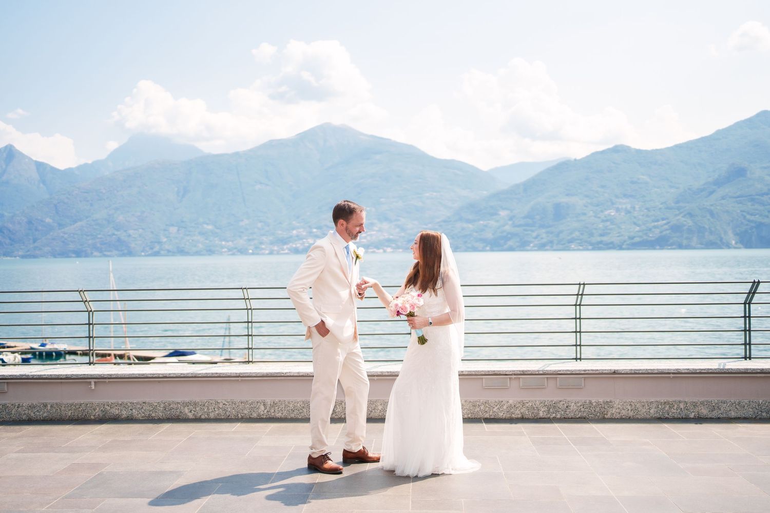 Bride and groom hold hands on a lakeside terrace with stunning mountain backdrop at an elegant destination wedding.