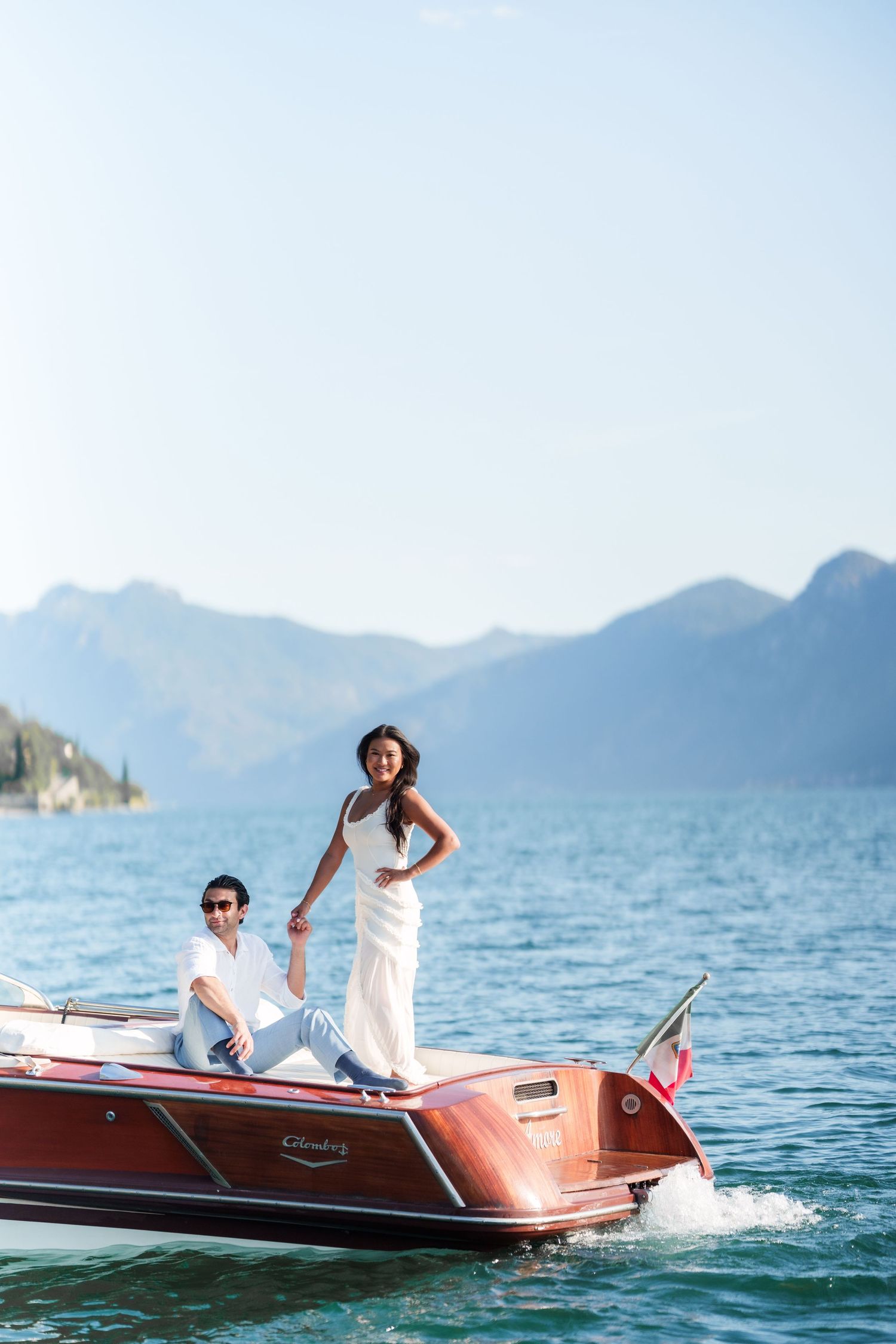 Couple in white on a classic wooden speedboat with mountain lake backdrop.