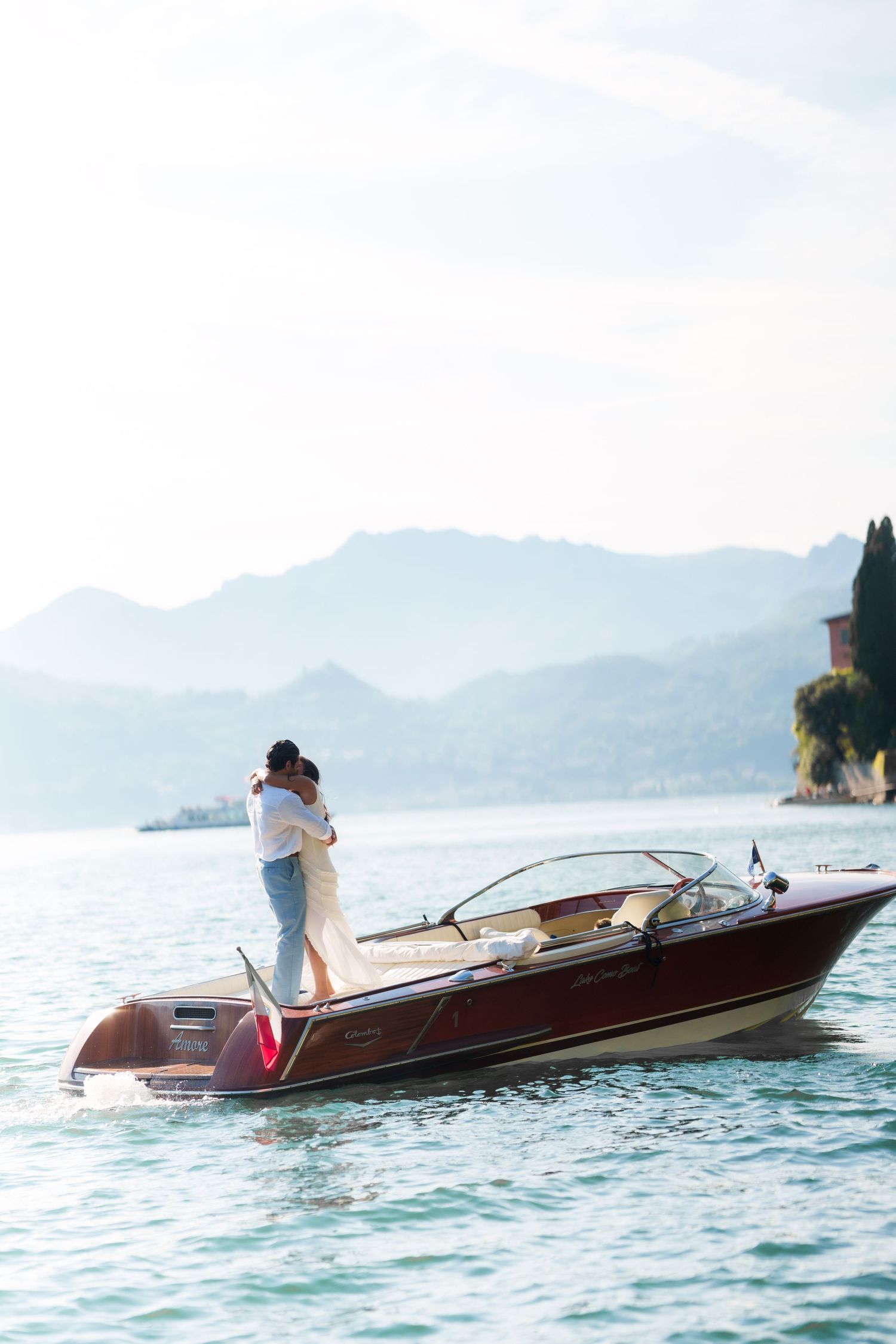 Couple embracing on a classic wooden speedboat on a serene alpine lake with misty mountains in the background.