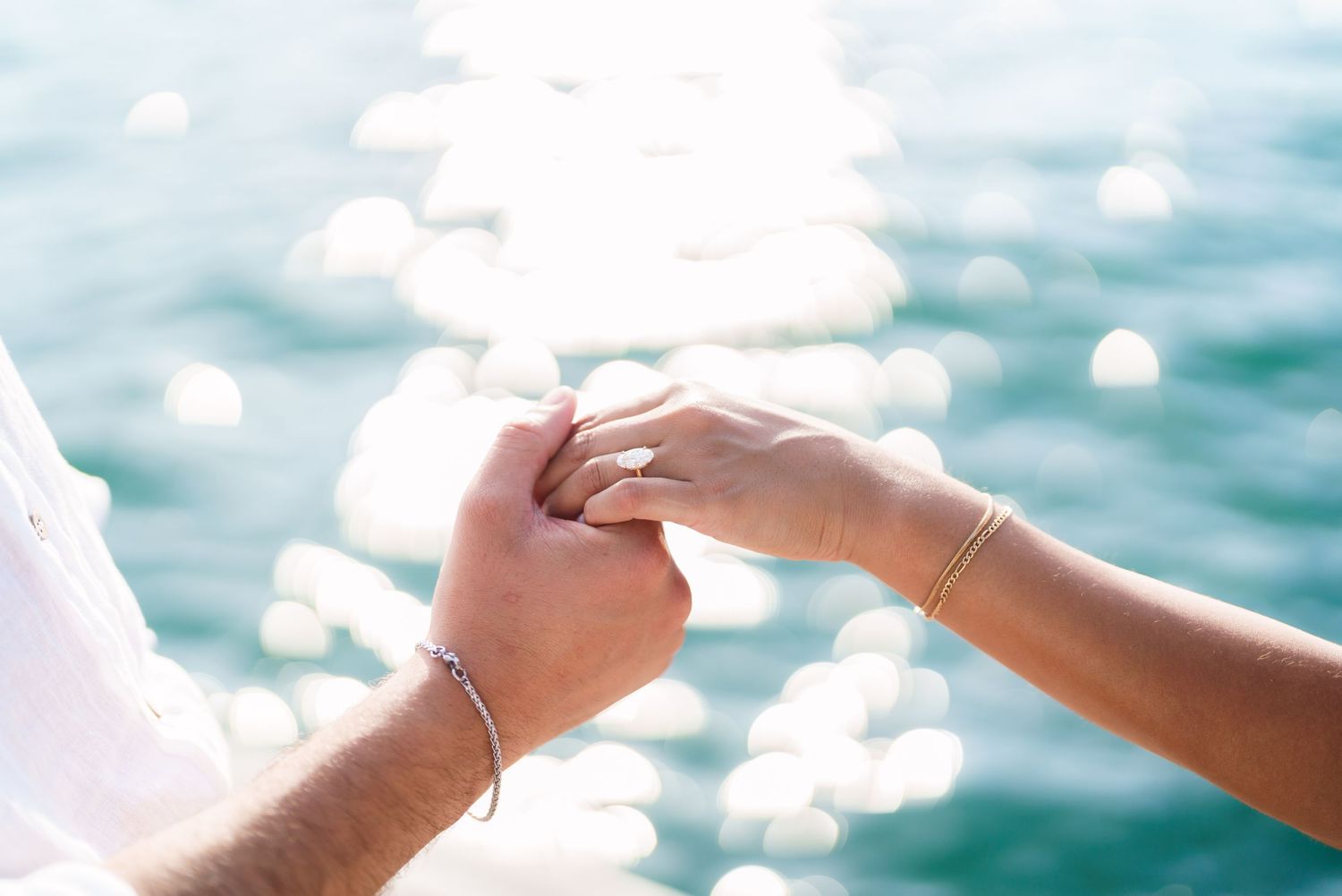 Couple holding hands tenderly over sparkling turquoise ocean water, woman wearing gold bracelet and engagement ring.