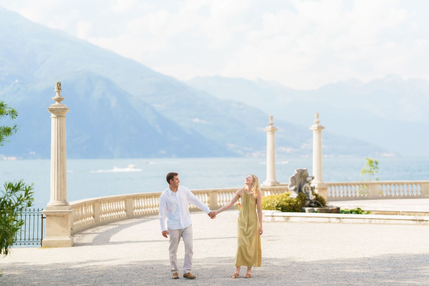 Couple holding hands on elegant terrace with classical columns overlooking Lake Como and mountains in Italy.