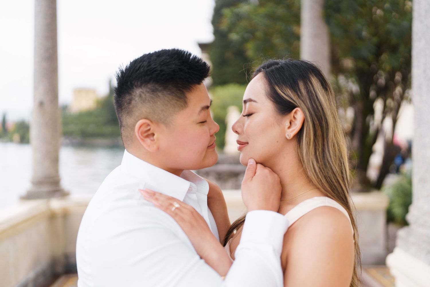 Couple sharing a romantic moment outdoors, man gently holding woman's face near lakeside columns.