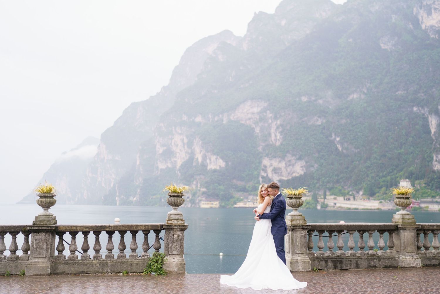 A couple shares a romantic moment on a misty balcony overlooking Lake Como with mountains in the background.