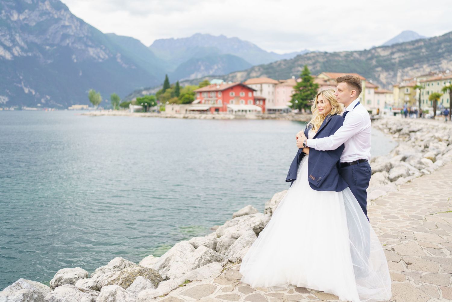 A couple embraces on a rocky shore with turquoise lake waters and majestic mountain peaks in the background.