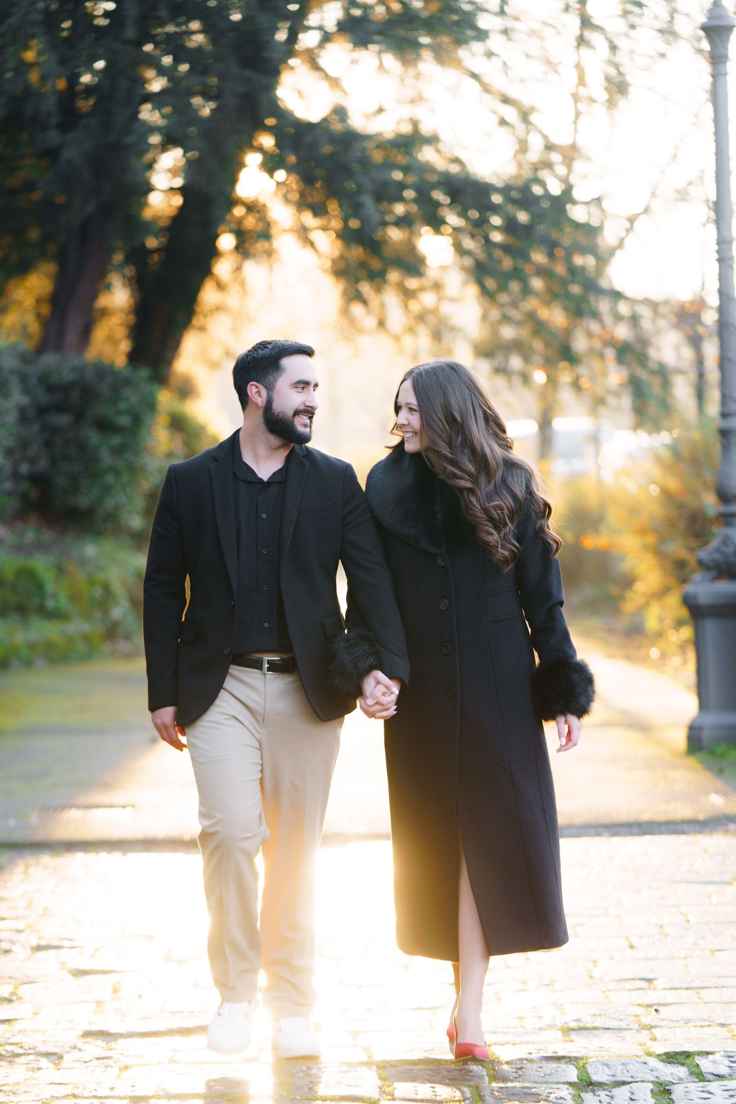 A couple in dark coats walks together on a sunlit path during golden hour, creating a romantic autumn atmosphere.
